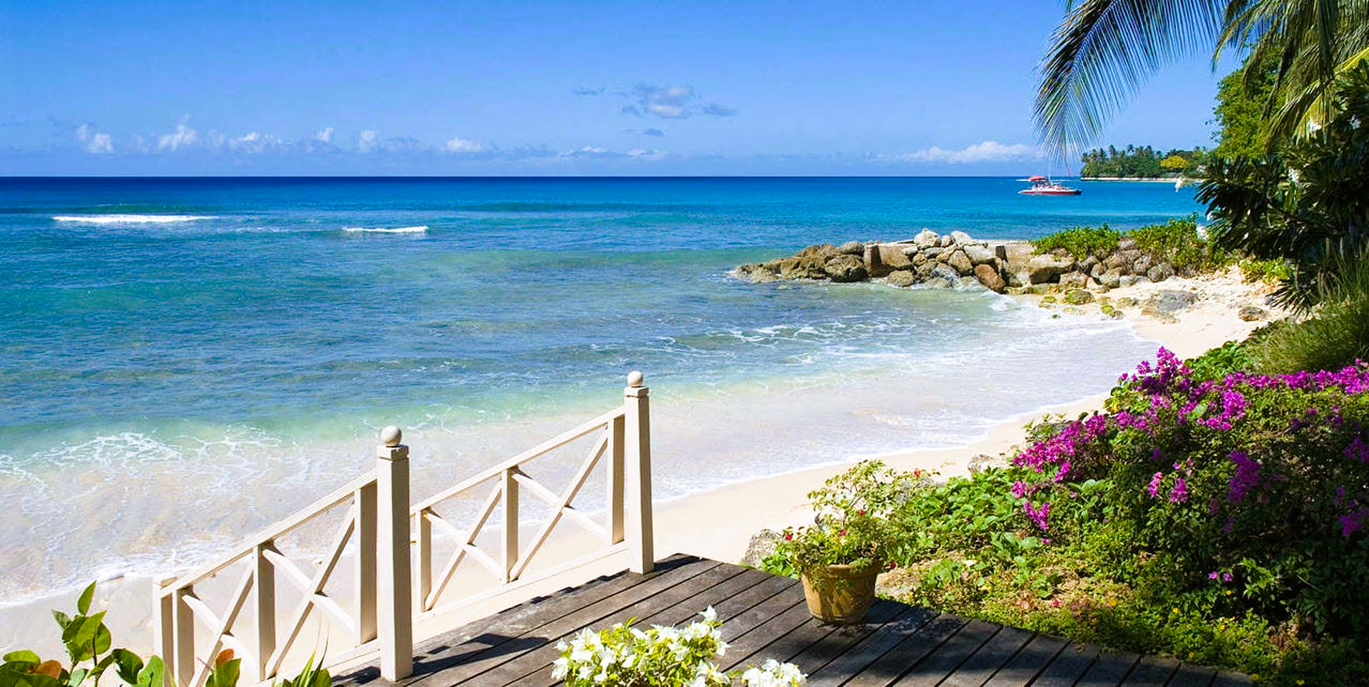 Beach scene with blue water, white sand, and flowering plants. A wooden deck and railing are in the foreground.