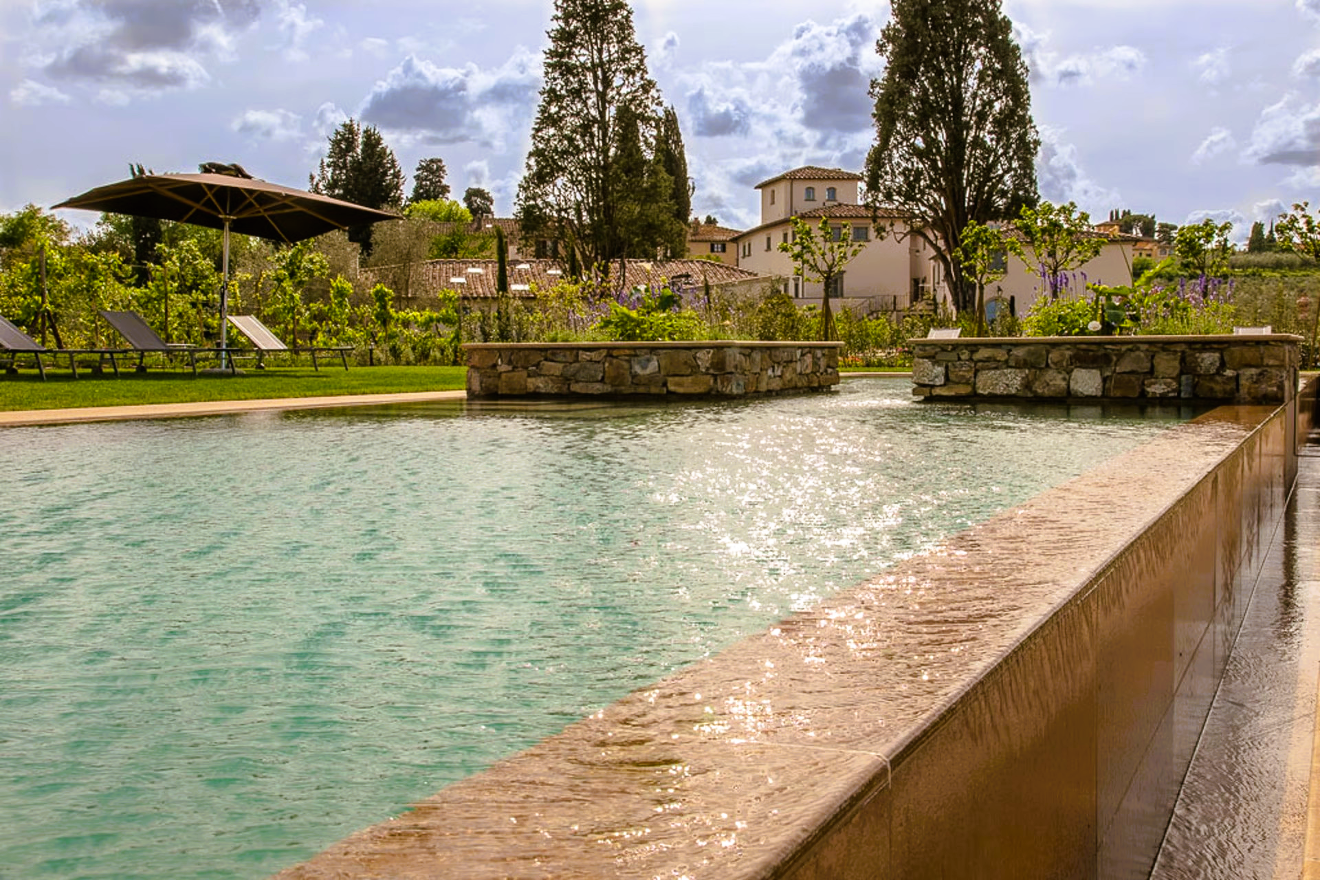 Pool with stone edge, flowing water, and Tuscan countryside backdrop.
