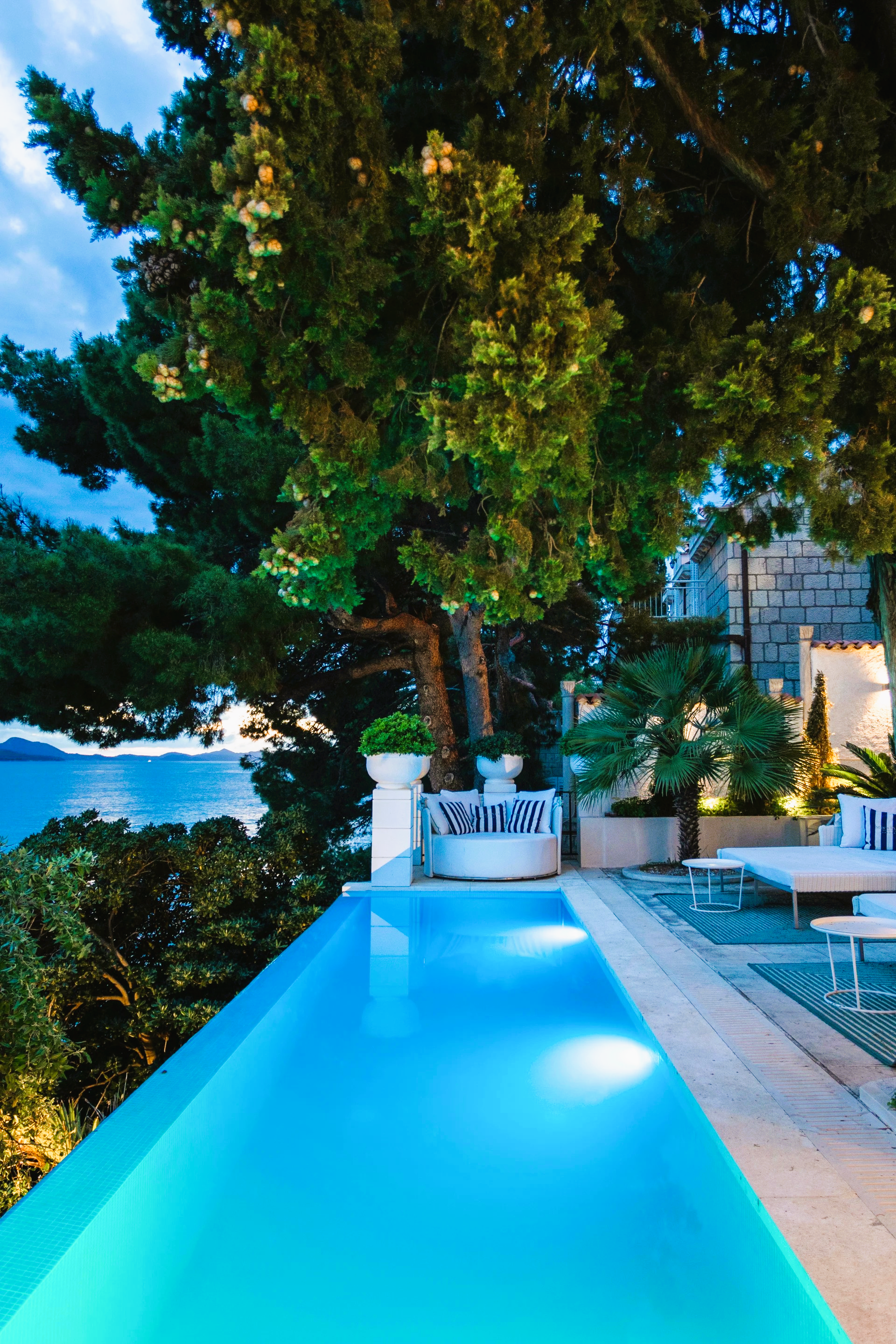 Infinity pool overlooking ocean, with trees, and lounge furniture.