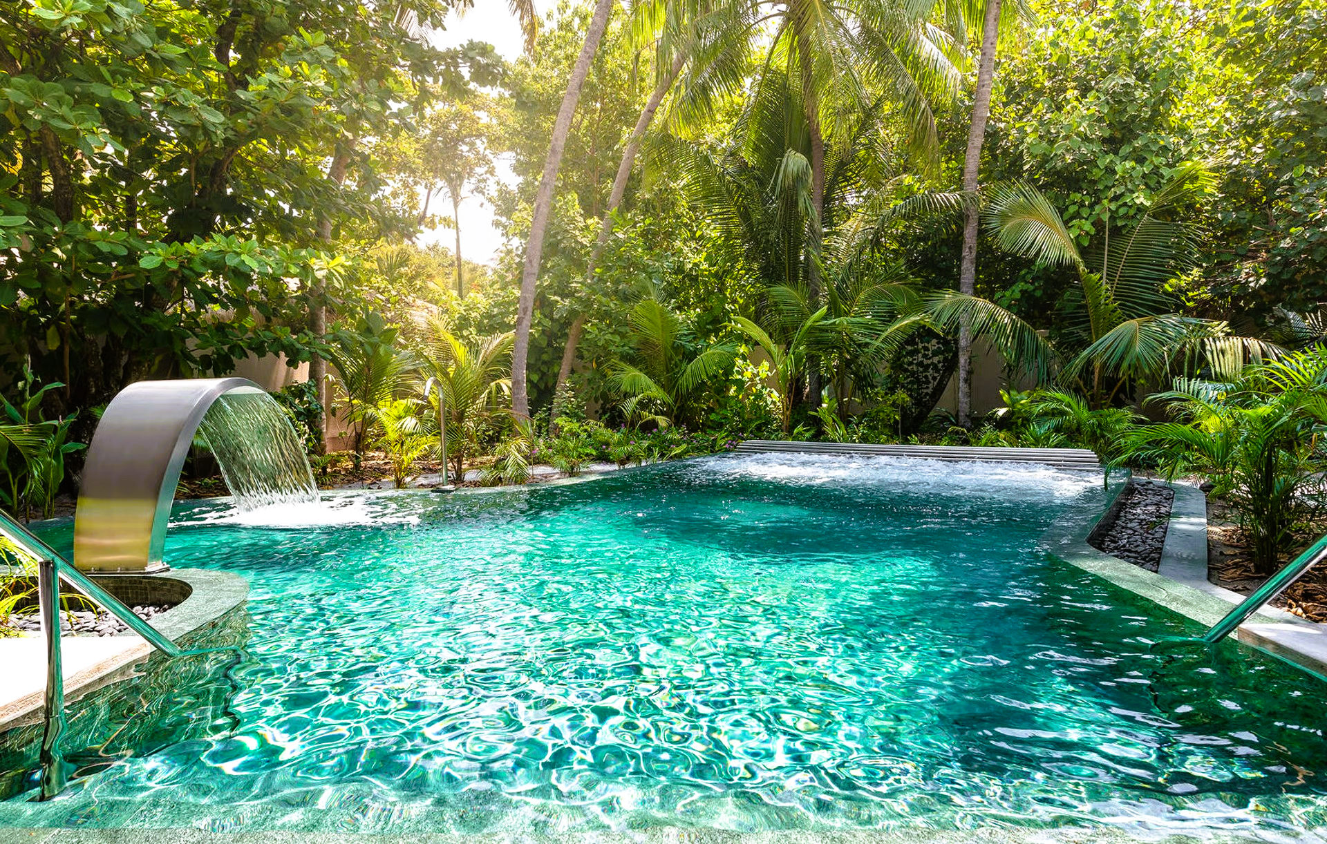 A turquoise pool with a waterfall feature surrounded by lush green tropical foliage.