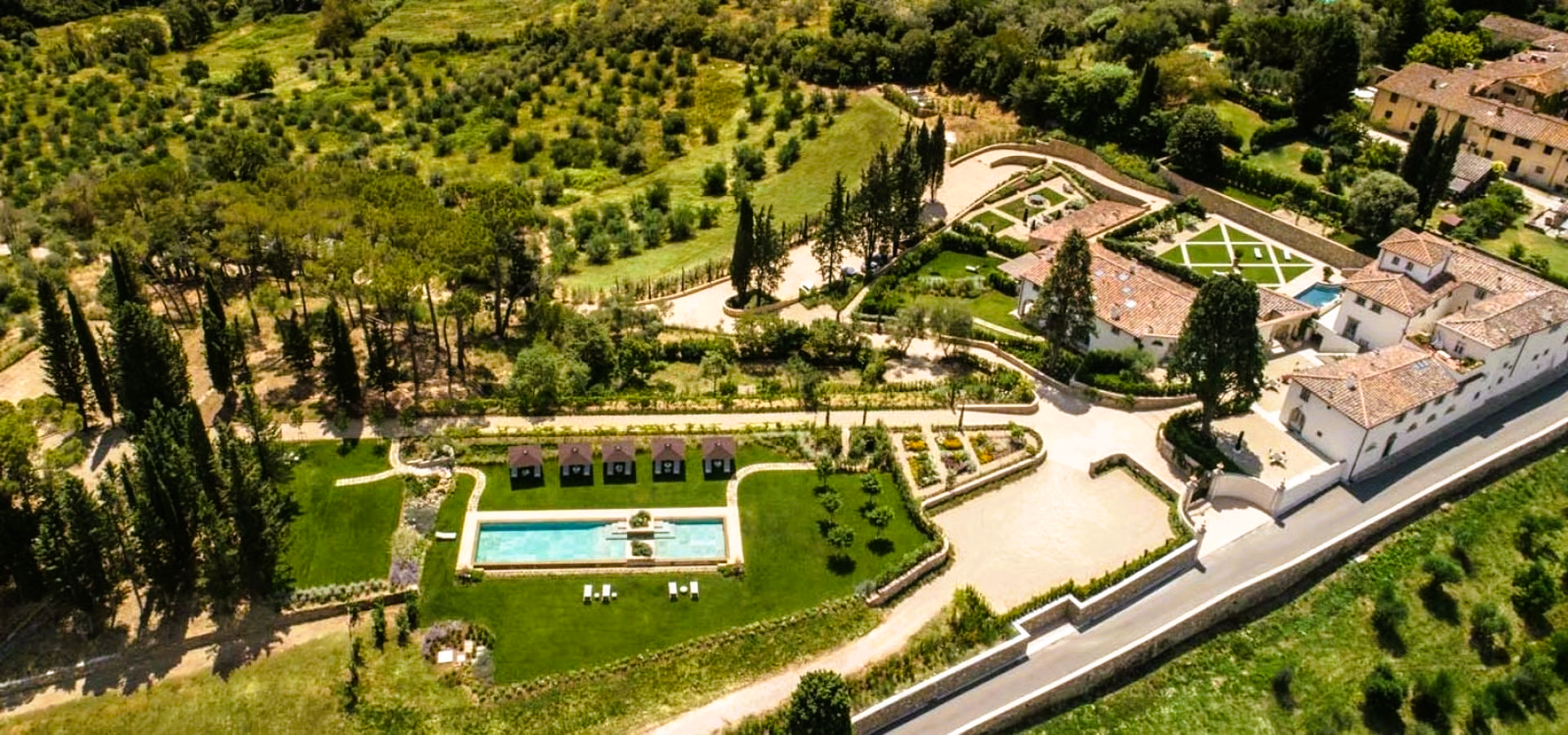 Aerial view of a luxury resort with a pool, gardens, and white buildings set in a green landscape.