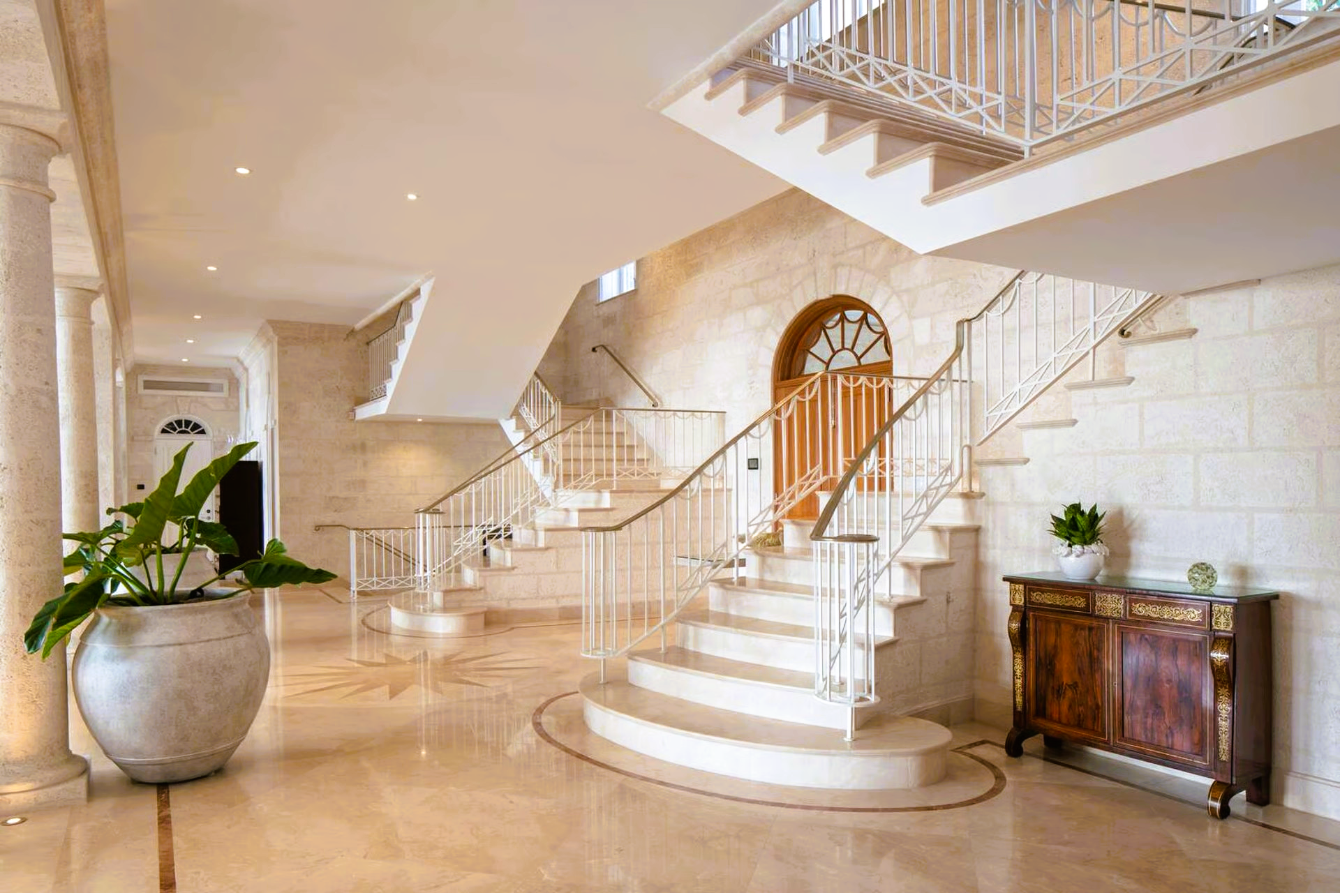 Grand, light-filled entryway with a curved staircase, stone walls, and a decorative wooden cabinet.