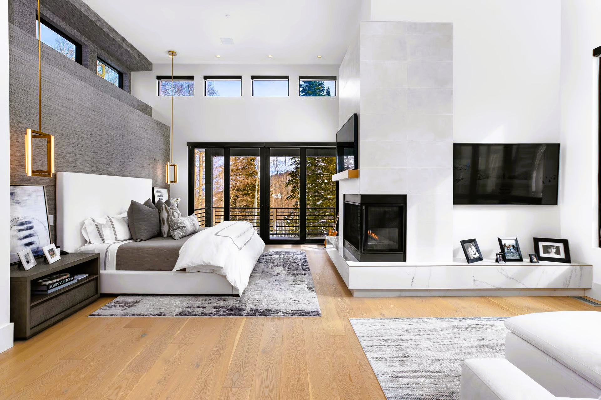 Spacious modern bedroom with a fireplace, bed, and large windows overlooking trees.