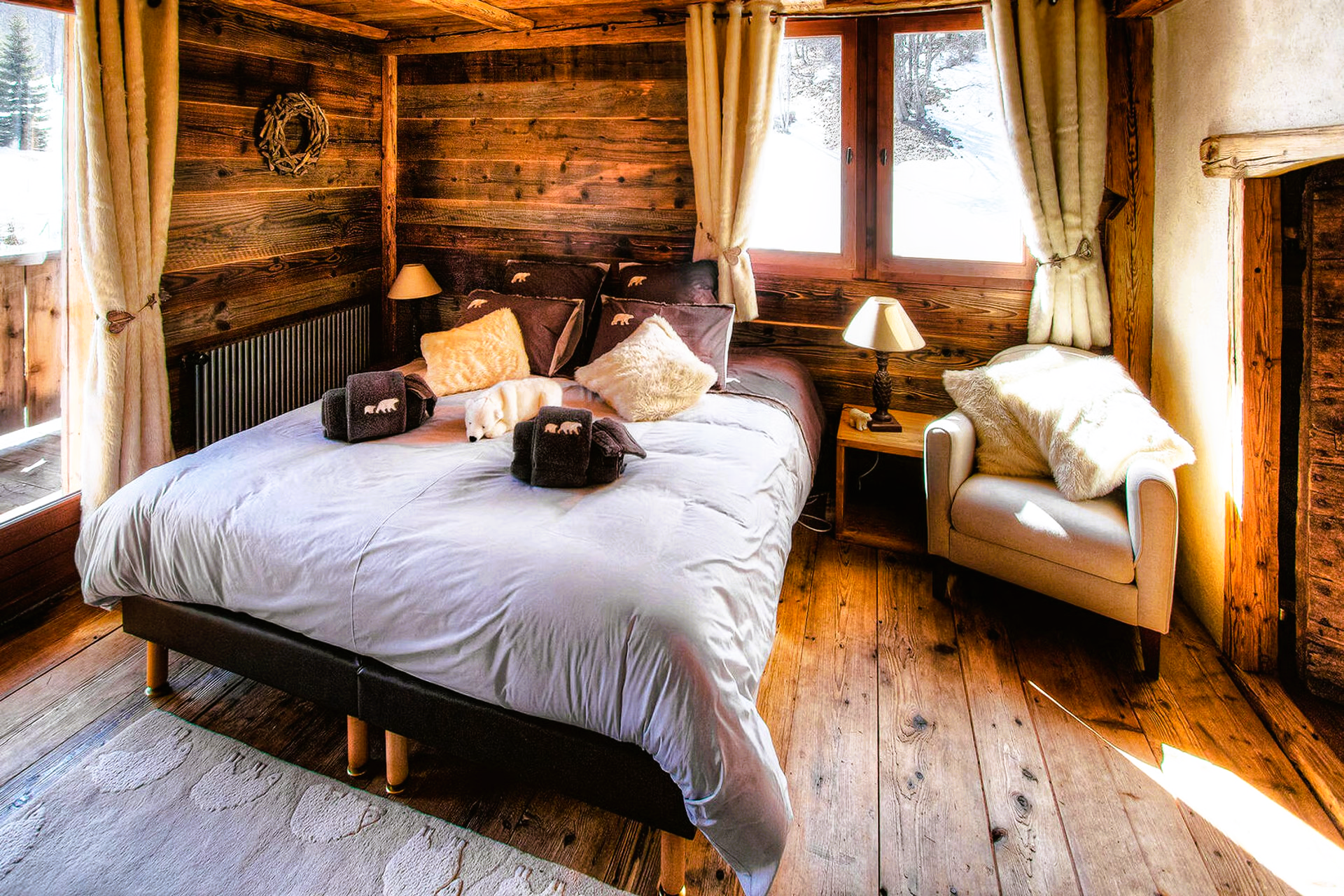 Cozy bedroom with wooden walls and floors. A large bed, armchair, window, and lamp fill the space.