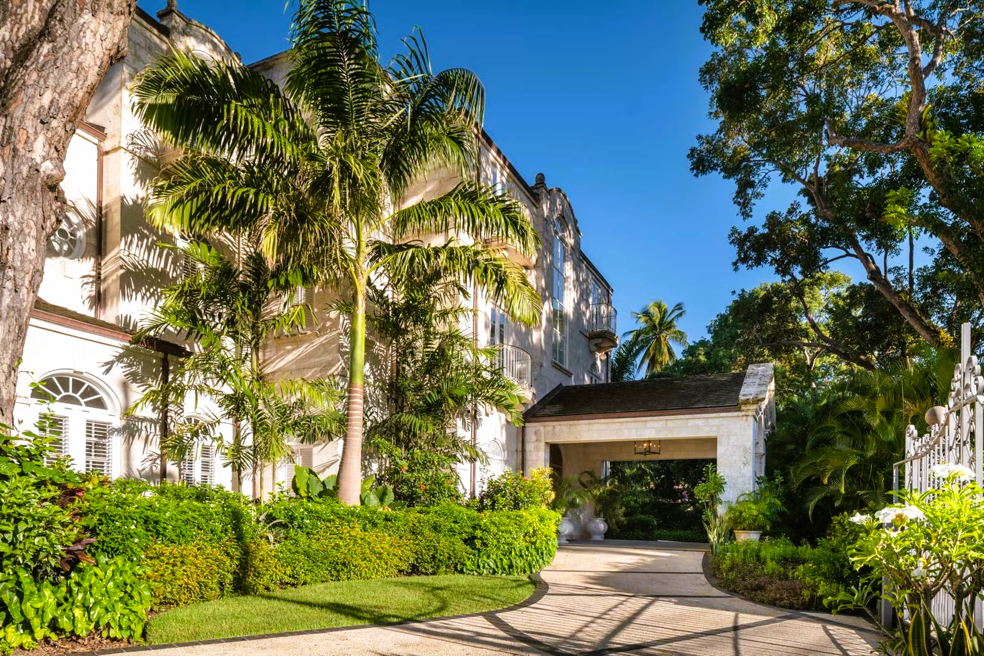 White building with arched windows, palm trees, and lush green landscaping under a blue sky.