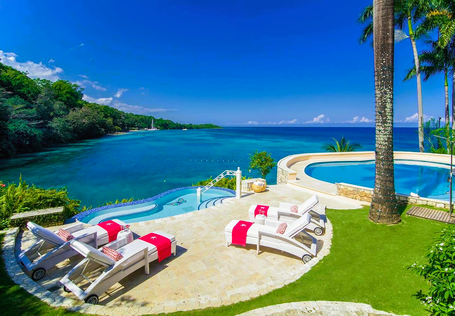 Lounge chairs on patio overlooking ocean and pool, bright blue sky, trees.