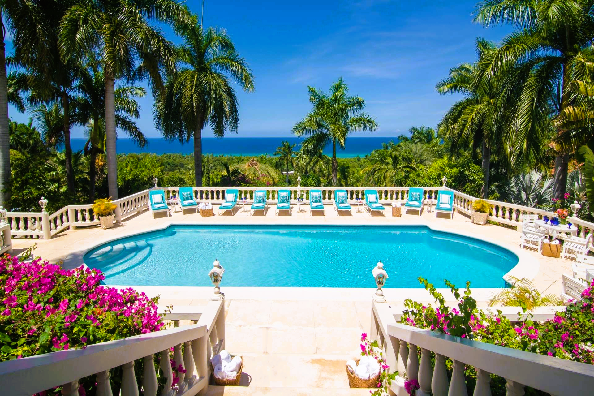 Swimming pool overlooking the ocean, surrounded by palm trees and flowers under a blue sky.