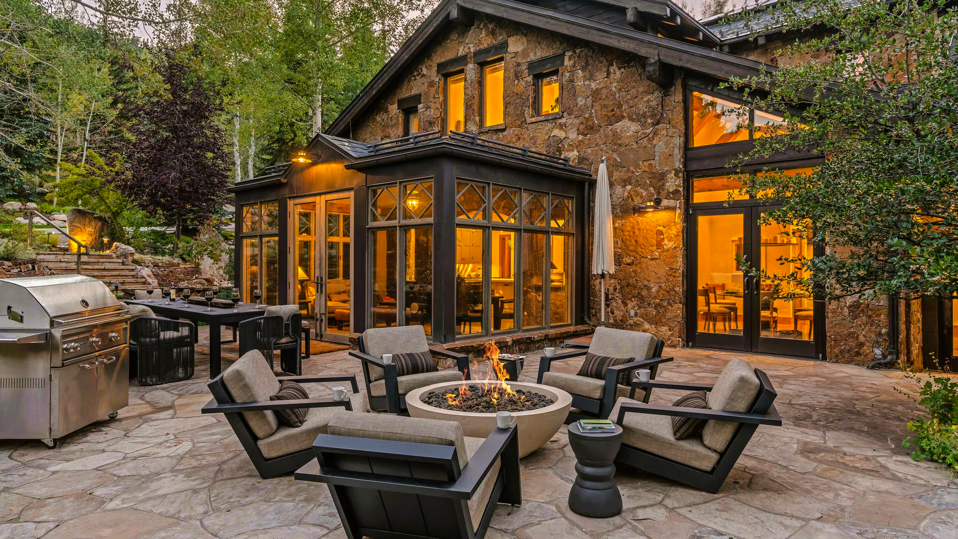 Patio with fire pit, seating, grill, and stone house with lit windows.