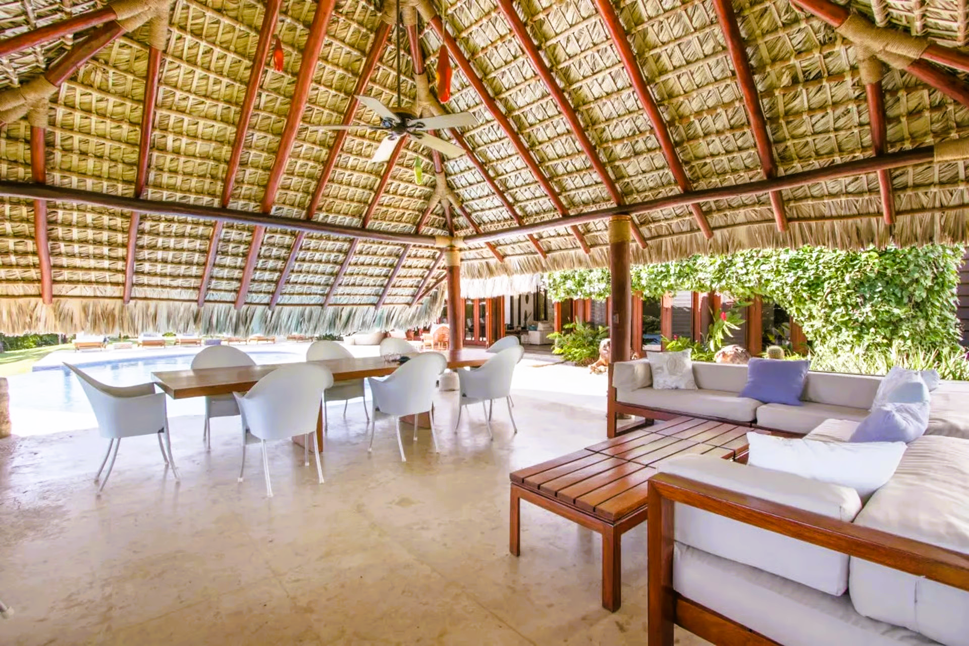 Open-air dining and lounge area with thatched roof, wooden furniture, and pool visible in the background.