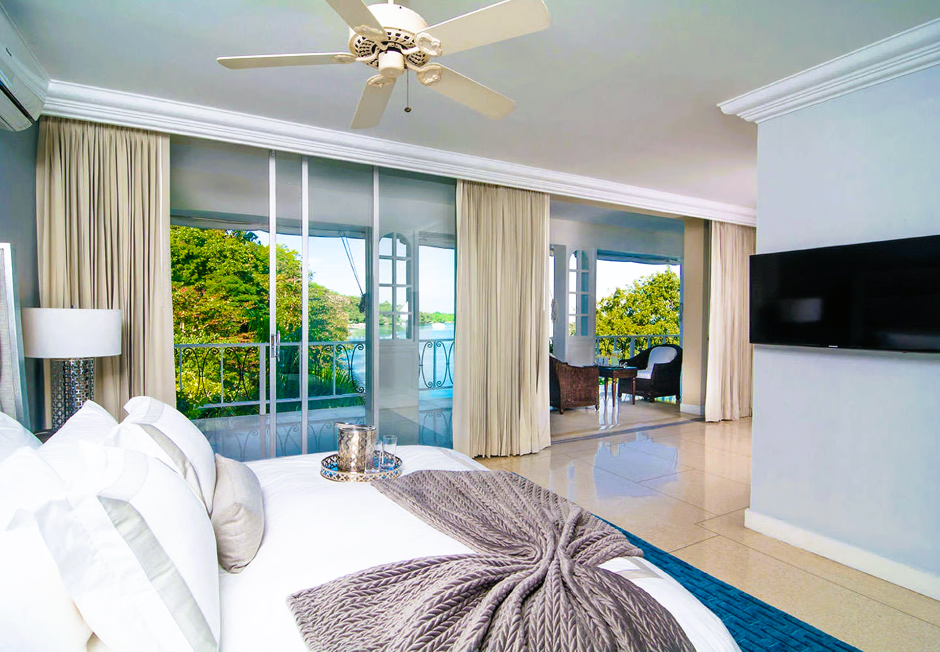 Bedroom with large windows overlooking greenery and water, a bed with pillows, a TV on the wall, and a ceiling fan.