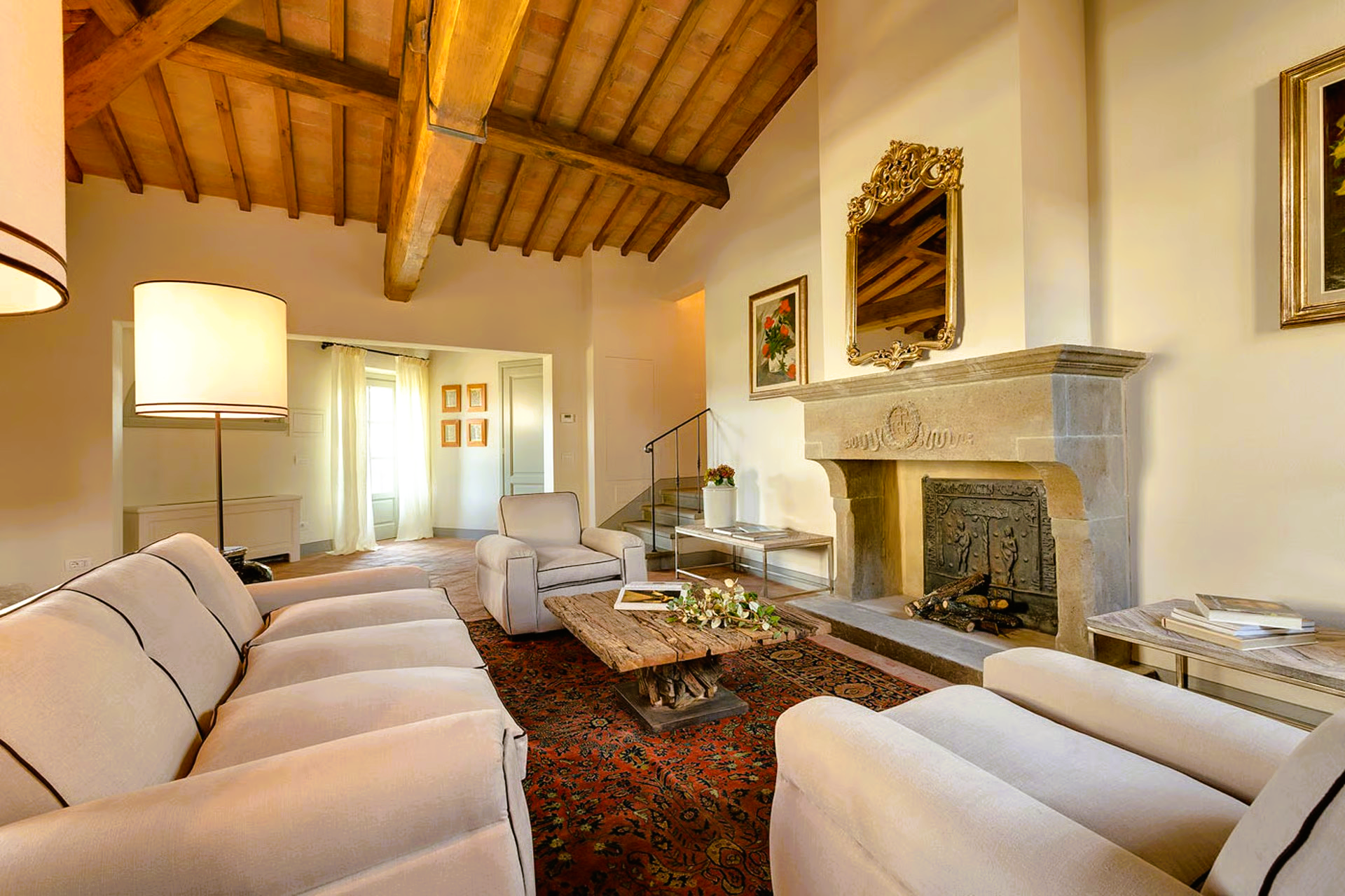 Cozy living room with cream-colored sofa, fireplace, and wooden ceiling beams in a sunny, Tuscan setting.