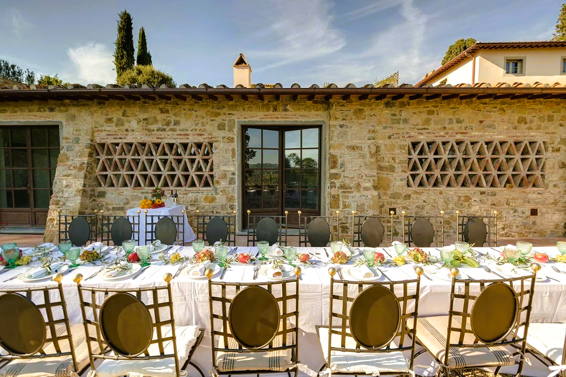Long table set for a meal outside a stone building. Chairs are gold with round backs.