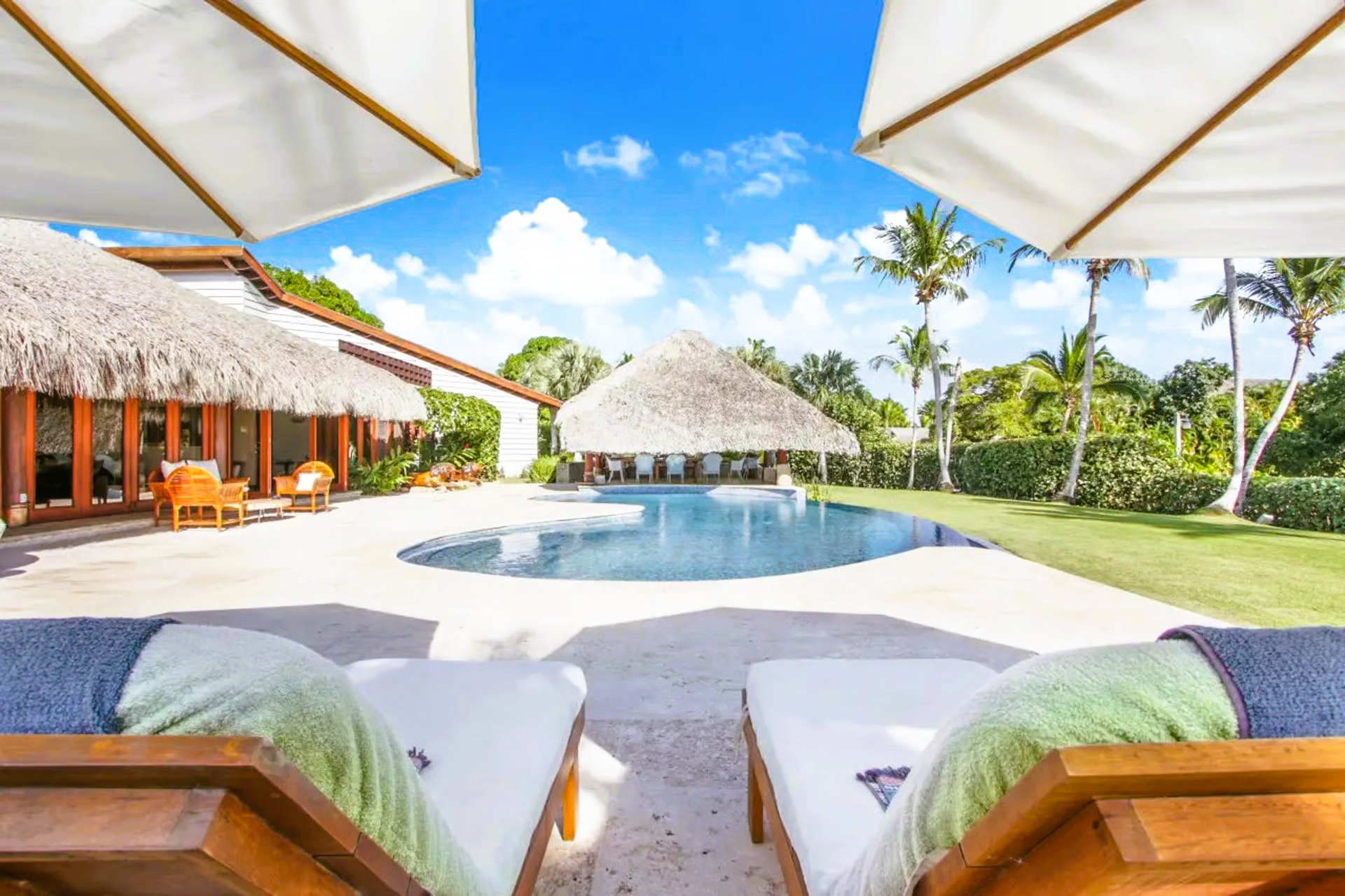 Two lounge chairs face a pool and thatched roof structures under a sunny blue sky with palm trees.