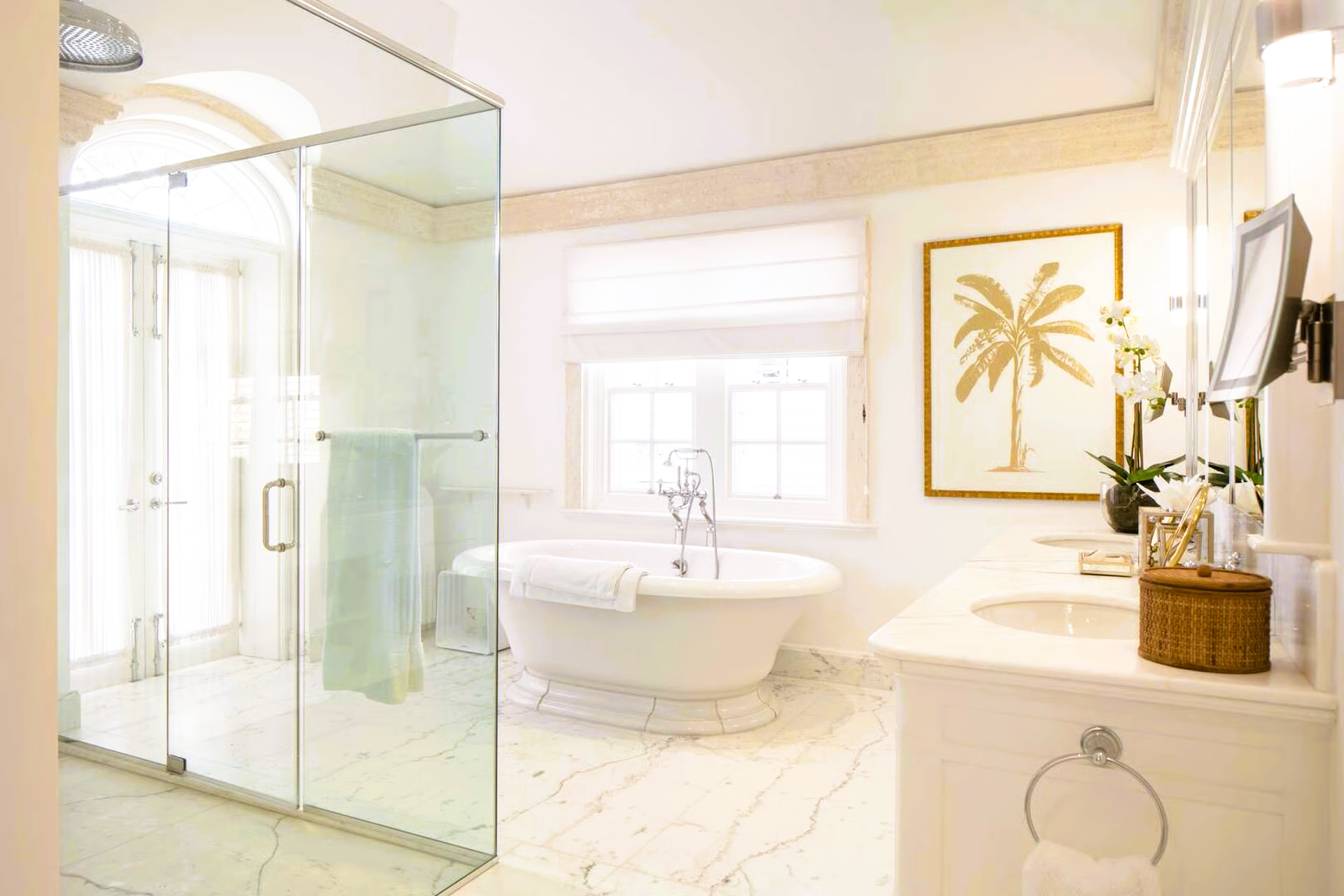 Bright, white bathroom with a glass shower, freestanding tub, and framed gold palm tree artwork.