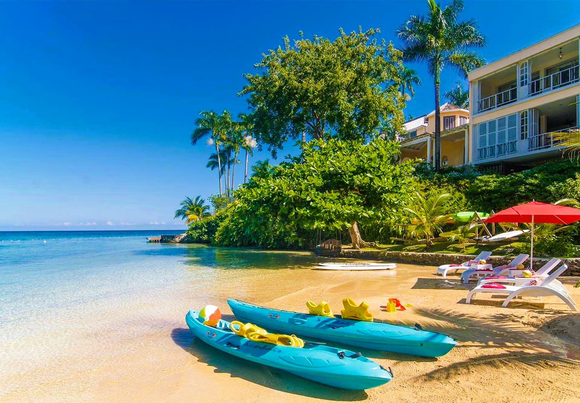 Beach with turquoise kayaks, clear water, and a resort under a bright blue sky.