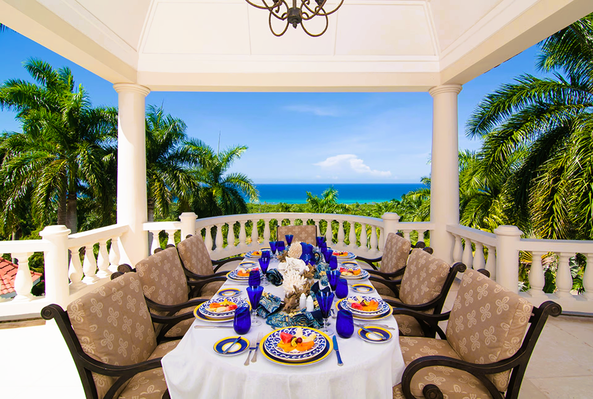 Outdoor dining table set for a meal with ocean view, white columns, and palm trees.