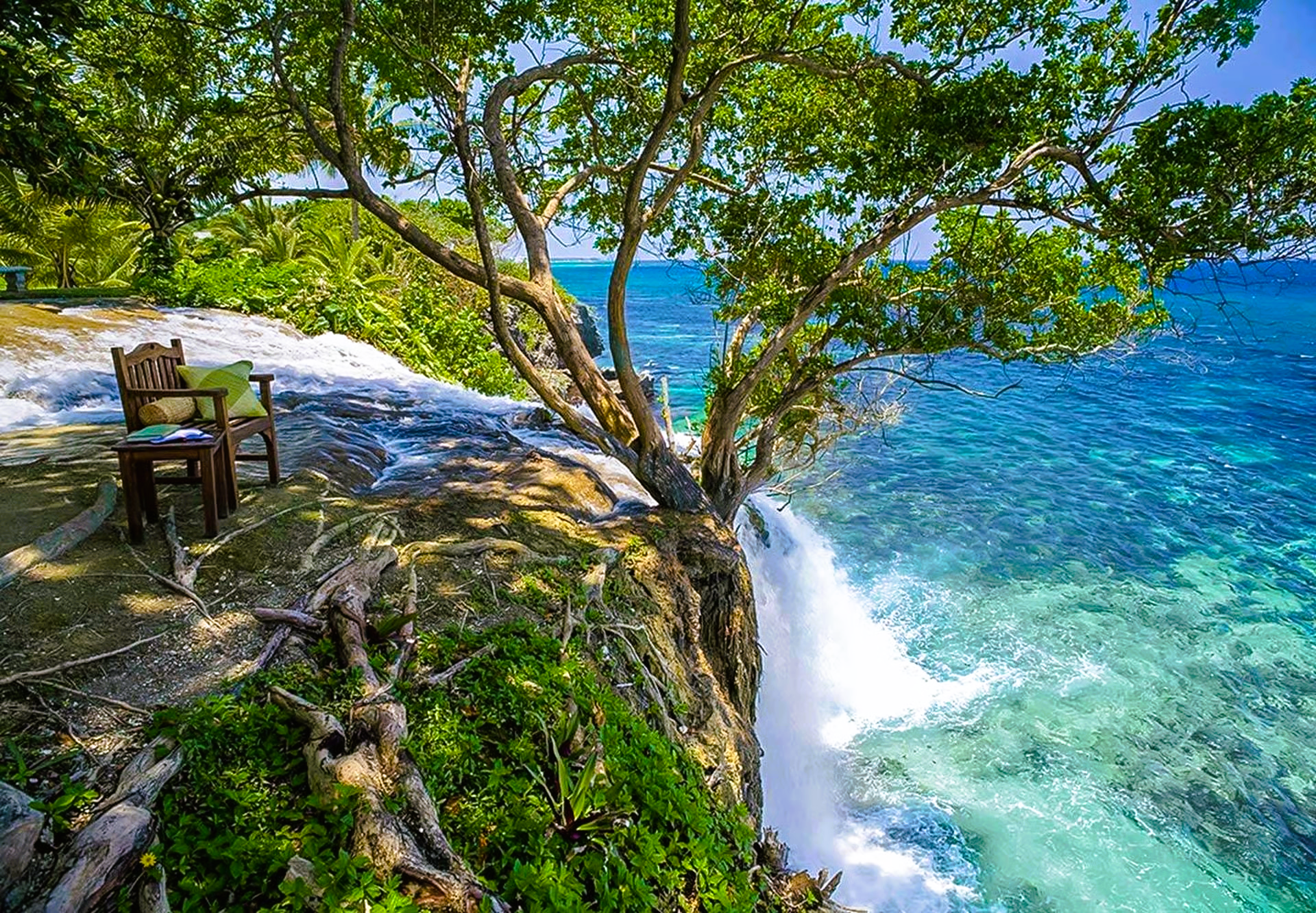 Cliffside view with a table and chairs under a tree, overlooking turquoise ocean water.