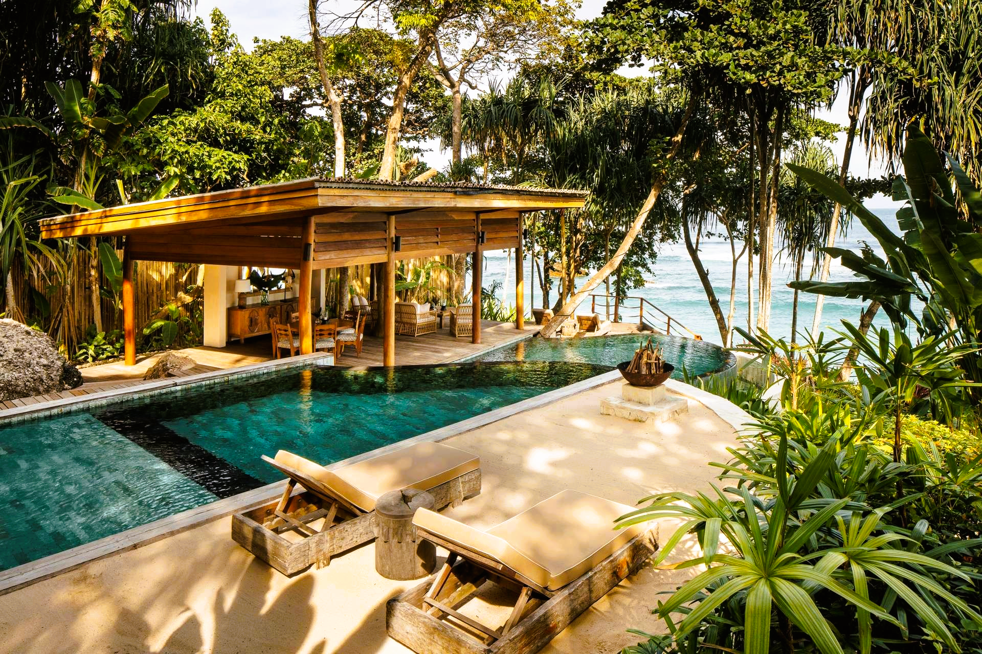 Tropical resort pool with ocean view, wooden cabana, lounge chairs, and lush greenery.