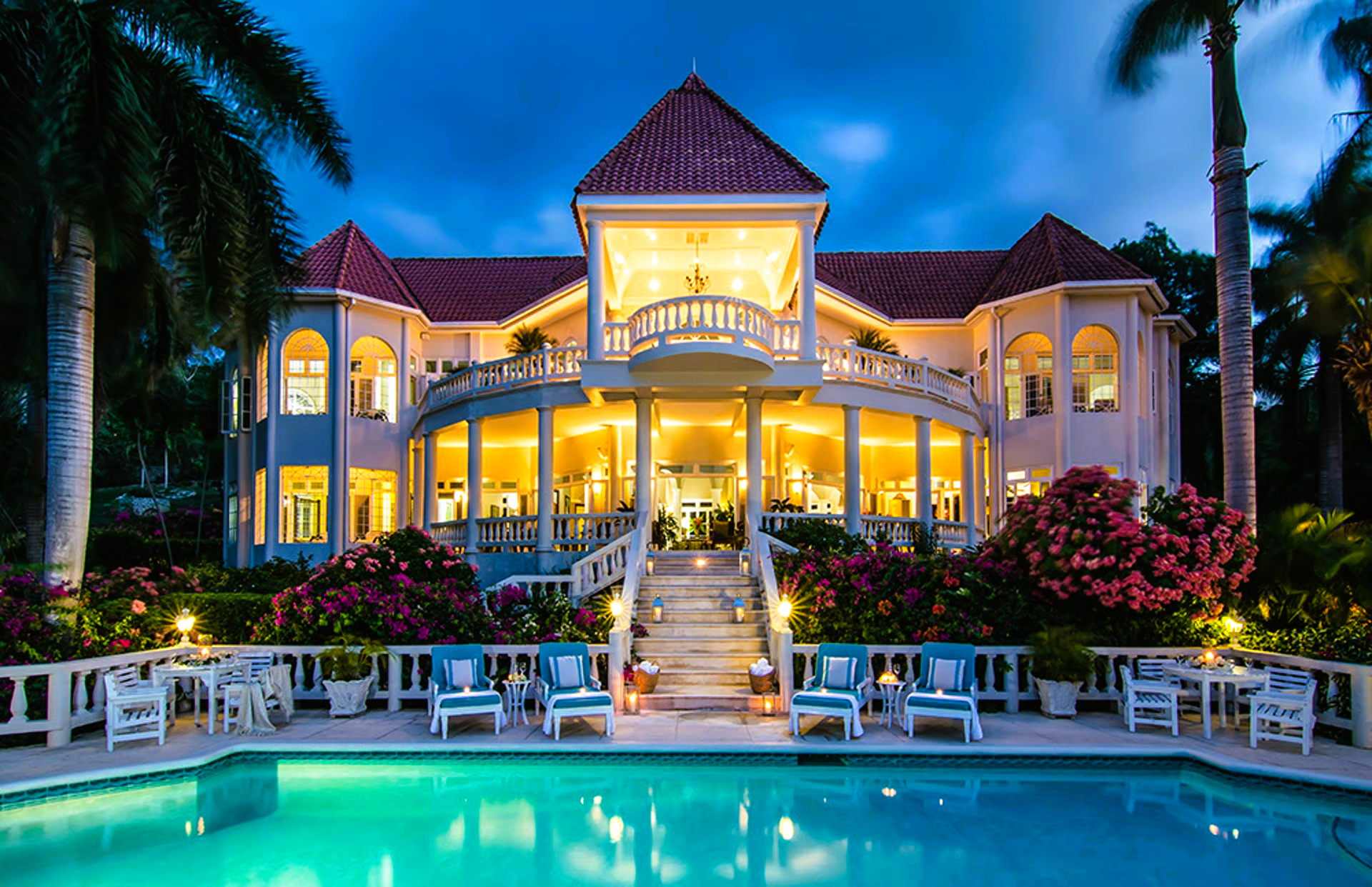 A luxurious, brightly lit mansion with a pool at dusk. White building with a red roof. Palm trees and blue lounge chairs.