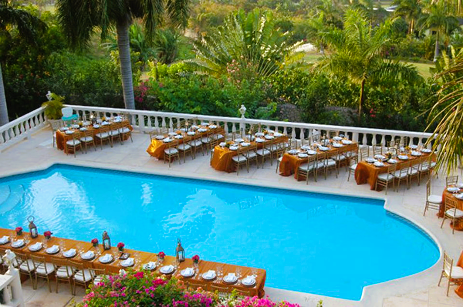 Tables set for an outdoor event beside a blue pool, surrounded by lush greenery and palm trees.
