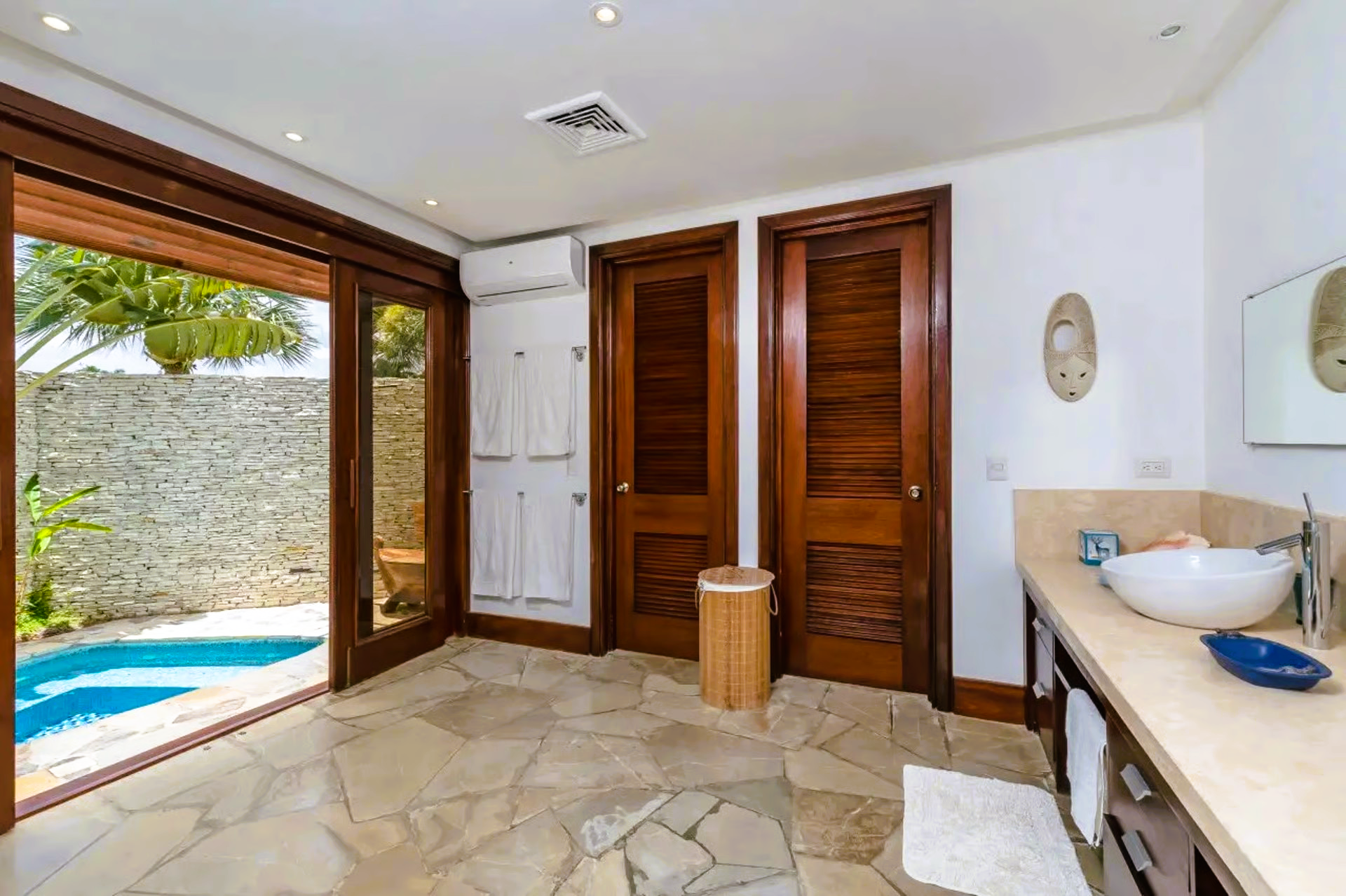 Bathroom with open doors to a small pool and stone wall. Features wooden doors, white towels, and a vanity.