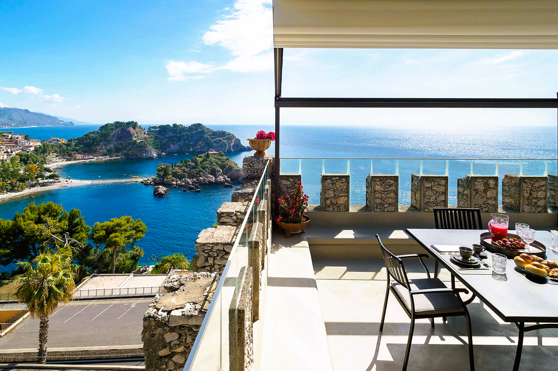 Balcony view of blue ocean, rocky coast, and a small town. Table set for a meal.