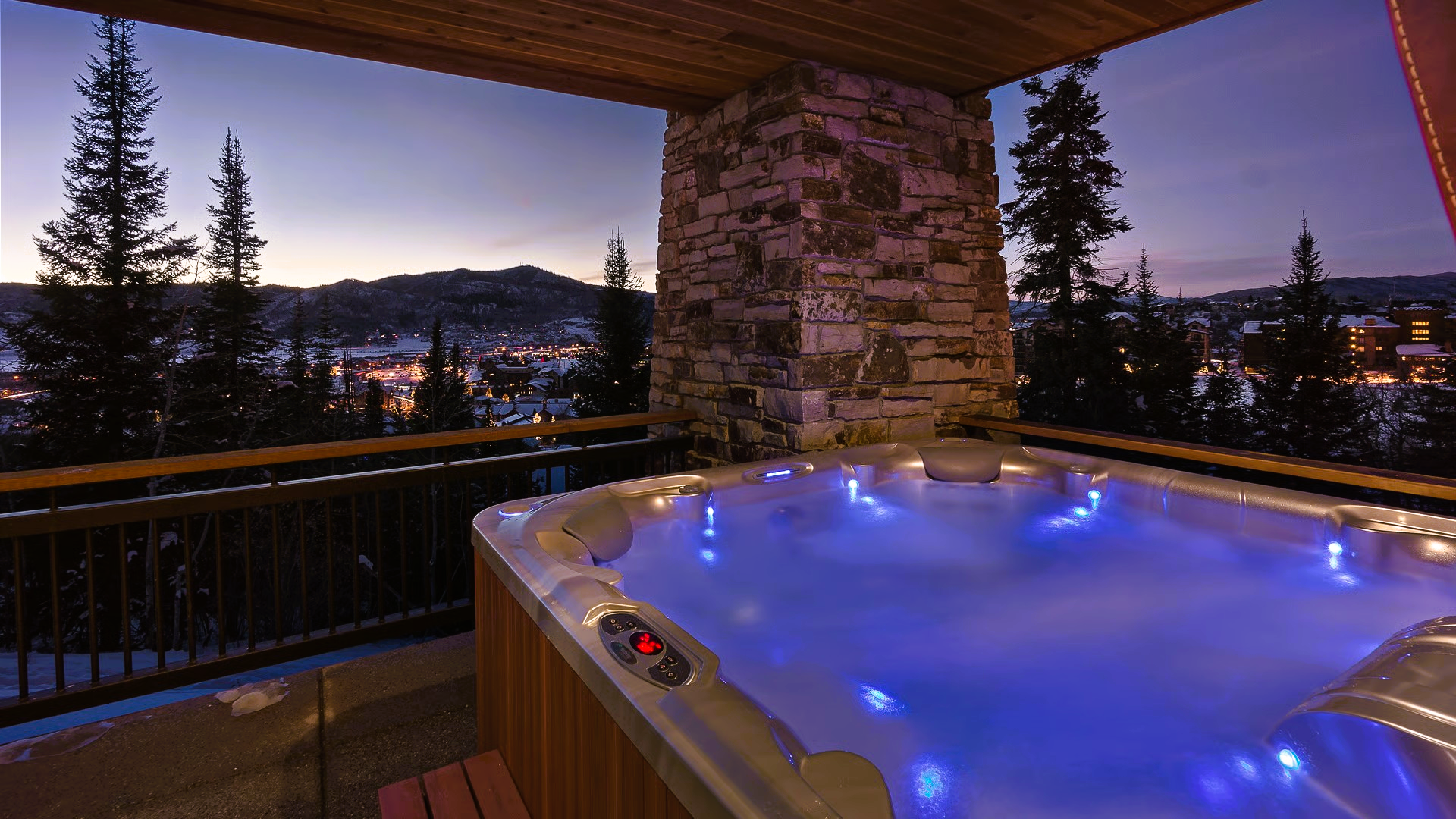 Hot tub on a balcony at dusk, overlooking a snowy town with lights; blue water, wooden accents.