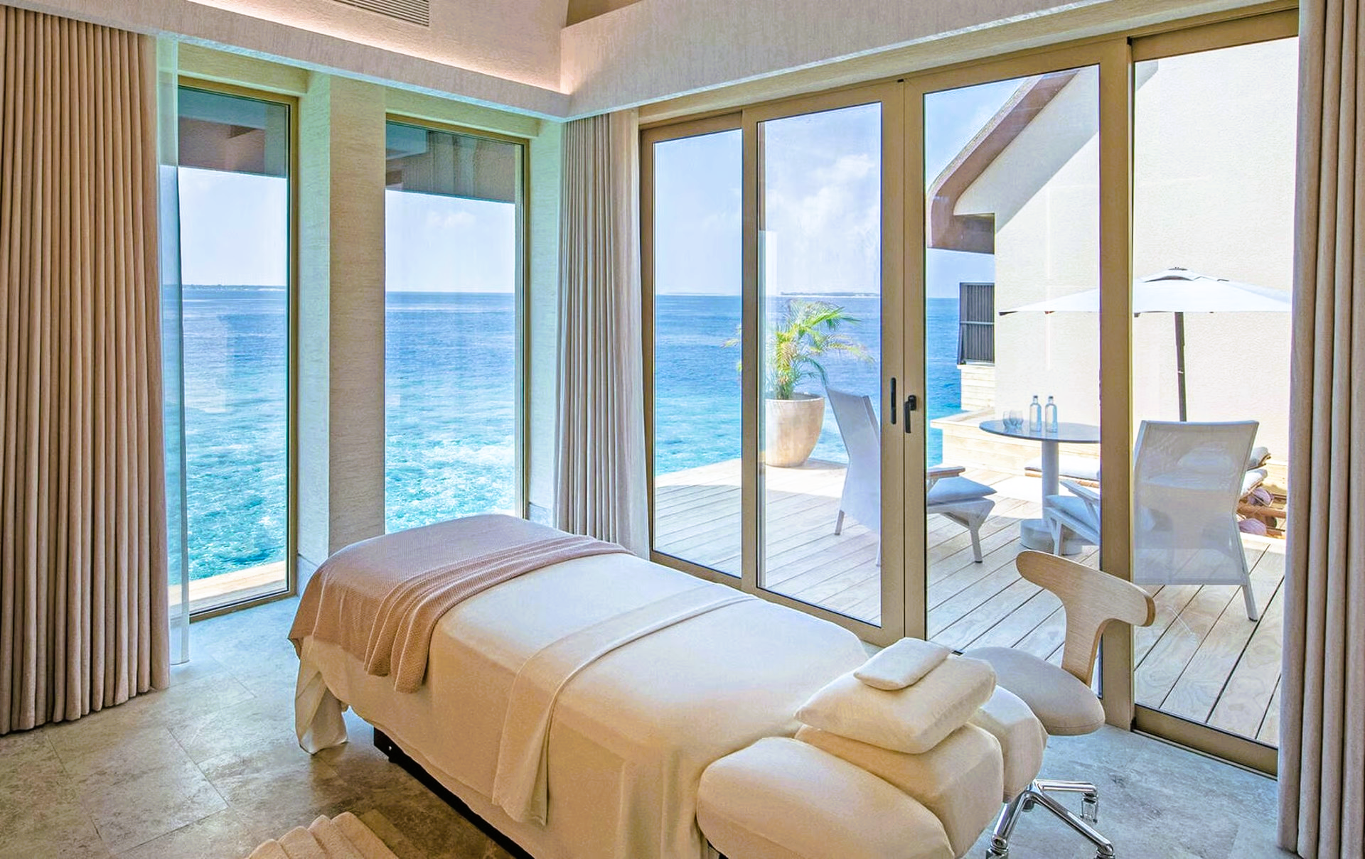 Massage table in a spa room with ocean view, glass doors, white decor and outdoor seating.