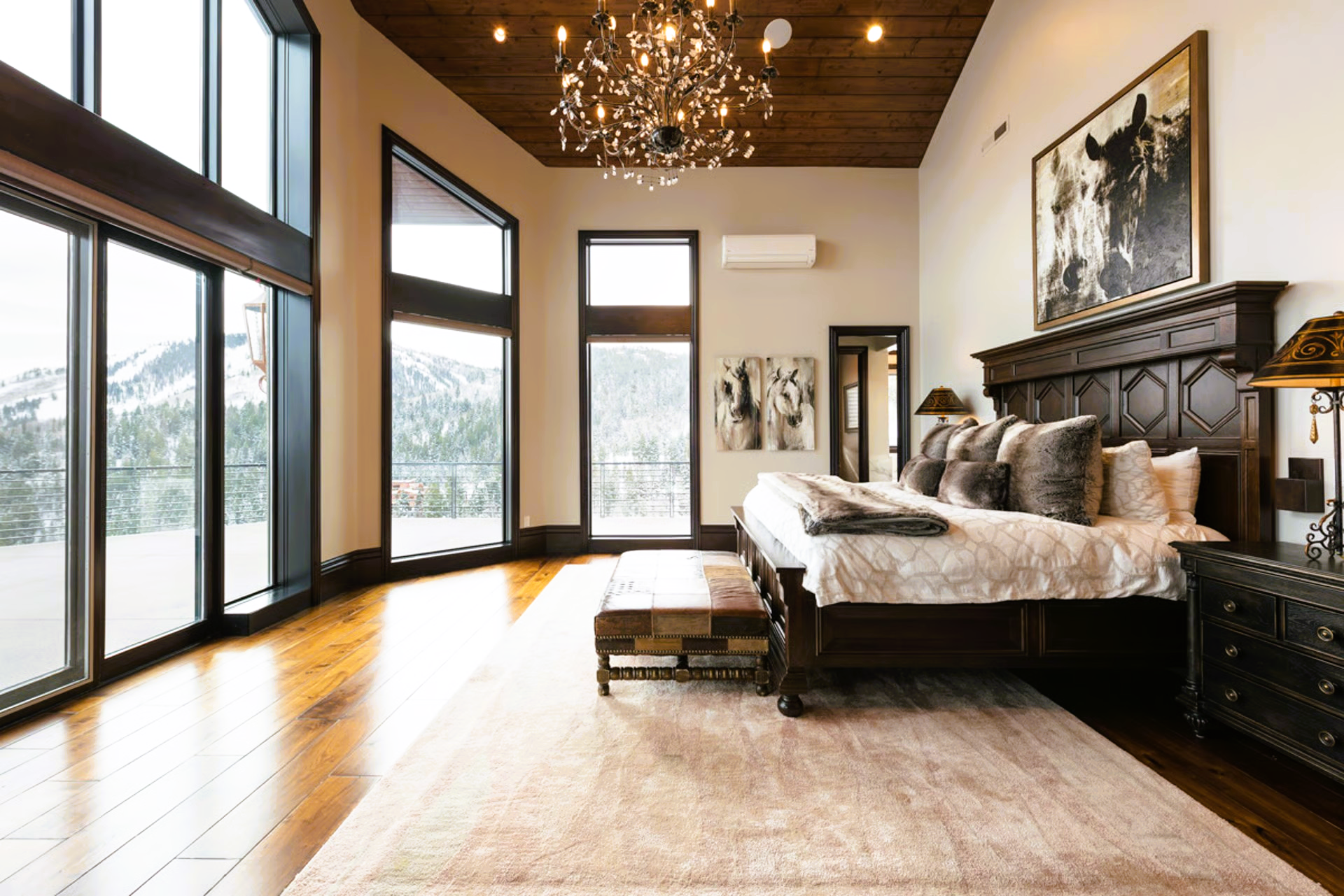 Spacious bedroom with large windows, wood ceiling, dark bedframe, and rug. Snow visible outside.