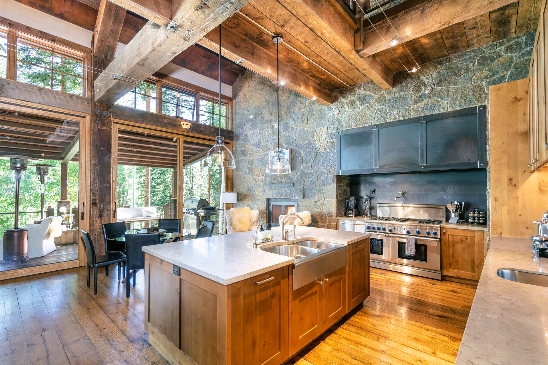 Kitchen with wooden cabinets, island, stainless steel appliances, and stone wall, overlooking a dining area and forest.