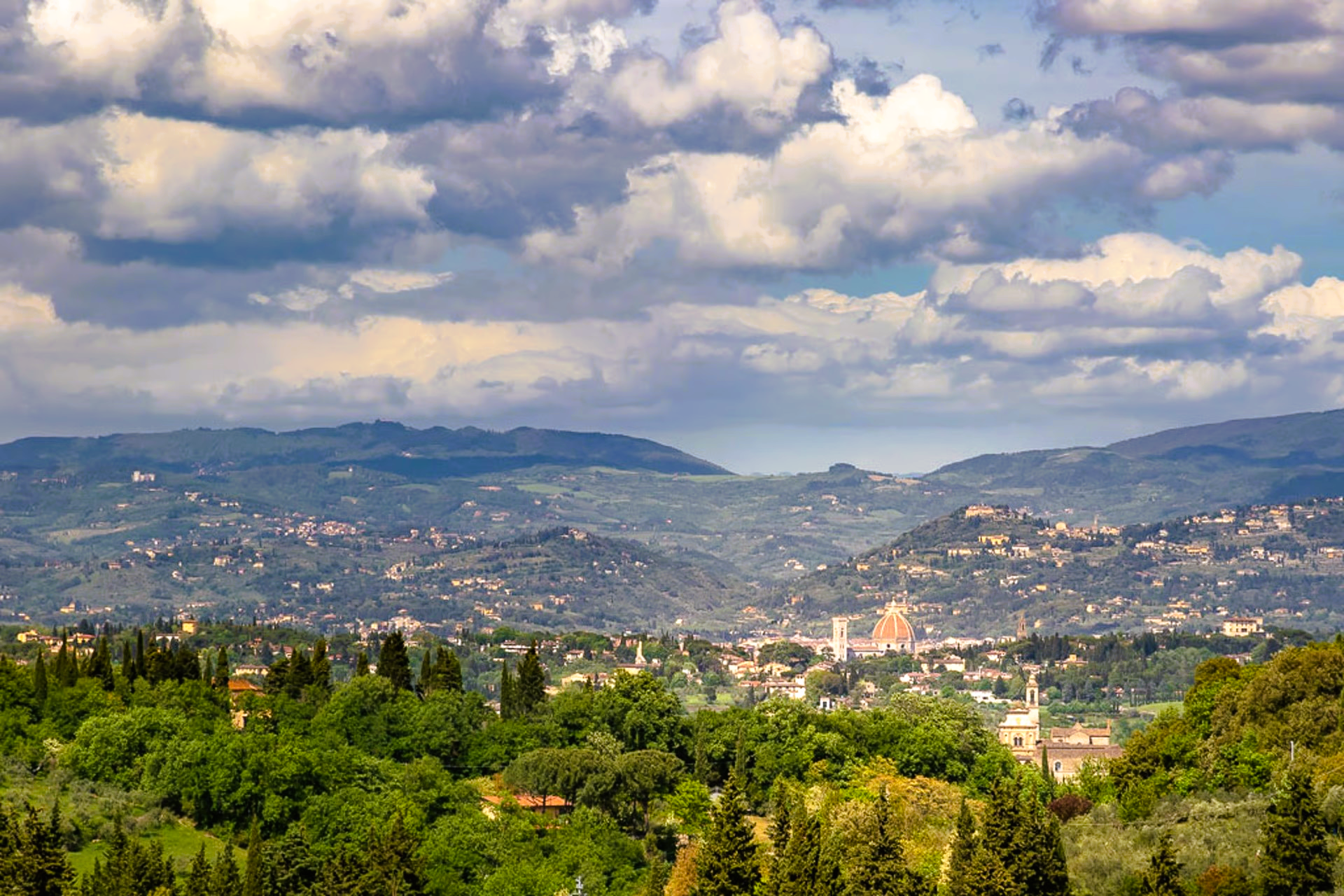 Hilly landscape with buildings and trees under a cloudy sky.