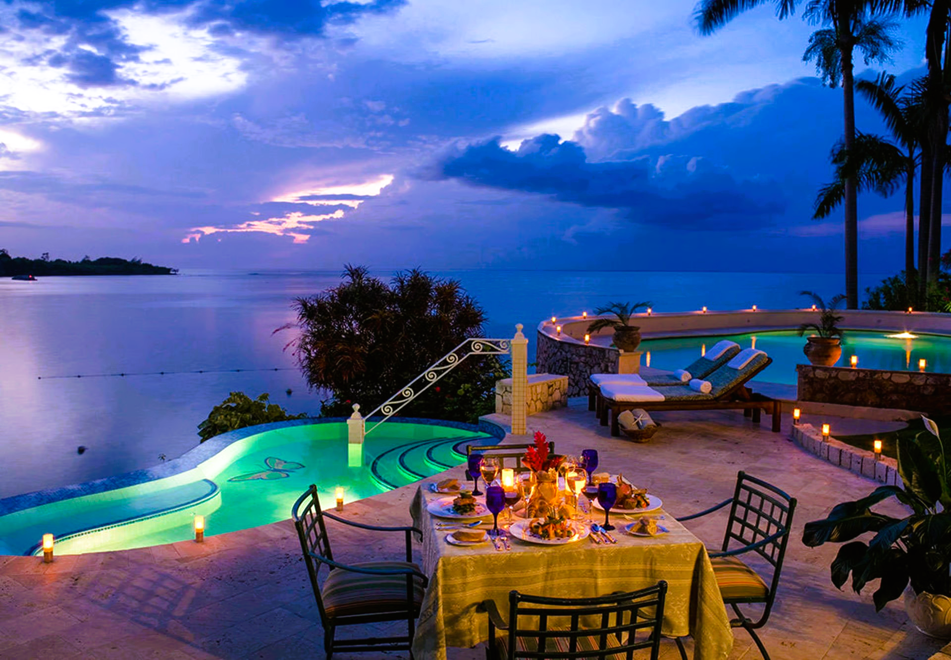 Table set for dinner by a pool overlooking the ocean at twilight.