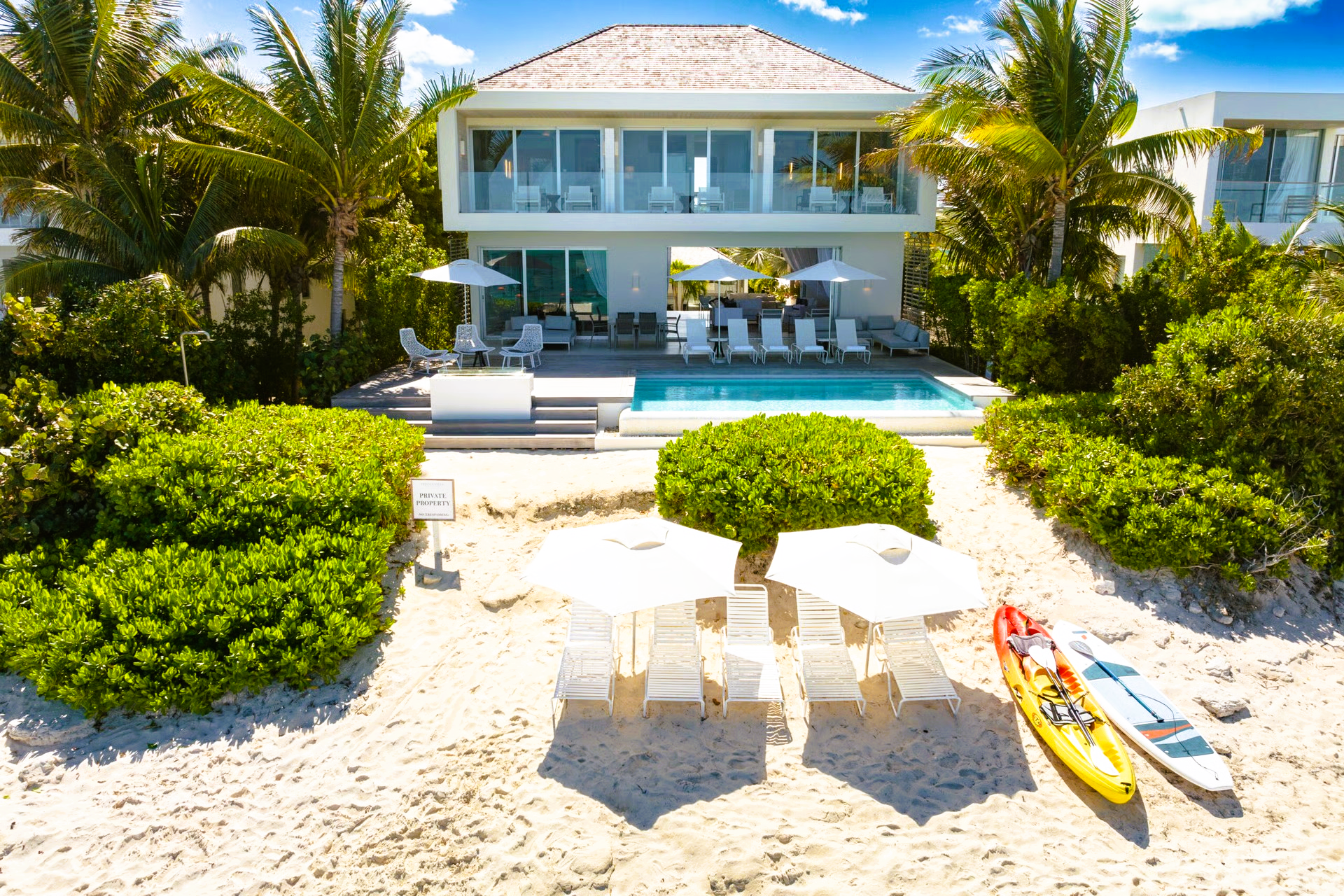 Beachfront villa with pool, umbrellas, kayaks; white building, palm trees, sunny day.