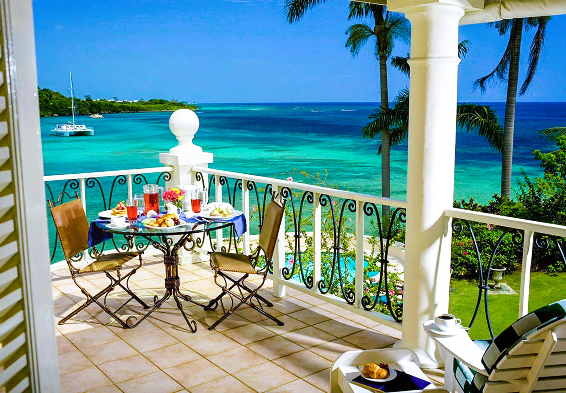 Balcony dining with ocean view: table set with food, turquoise water, palm trees.