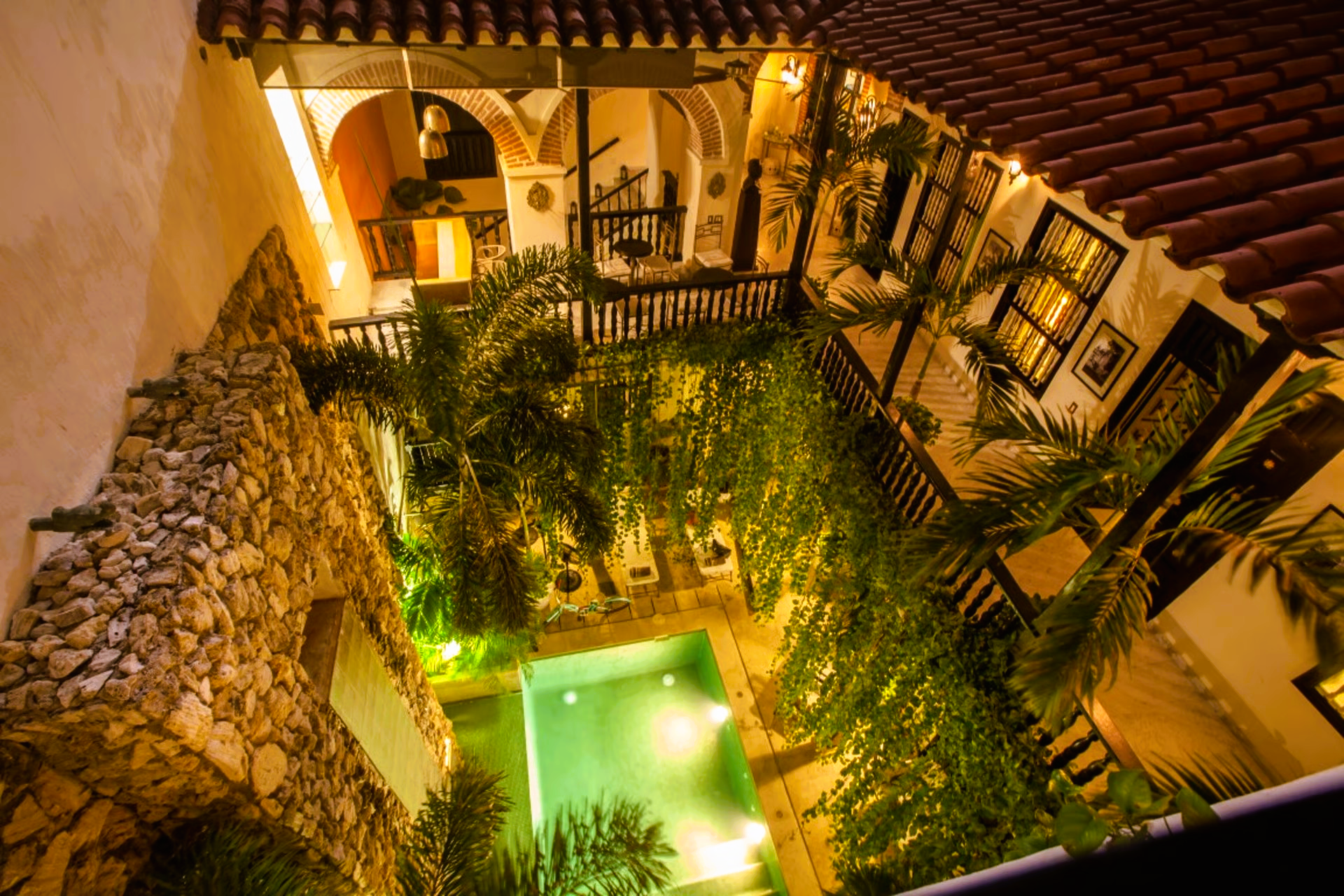 Courtyard with a pool surrounded by lush greenery and illuminated archways, at night.
