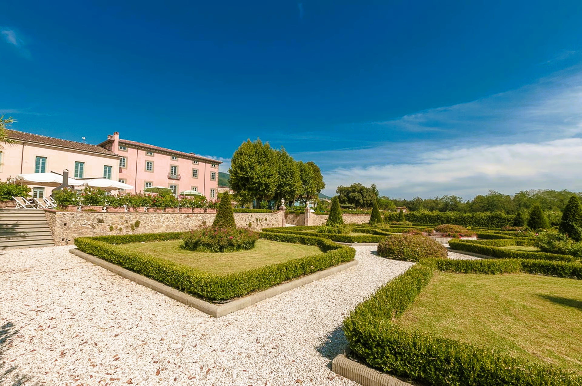 Formal garden with neatly trimmed hedges, gravel paths, and a pink building under a clear blue sky.