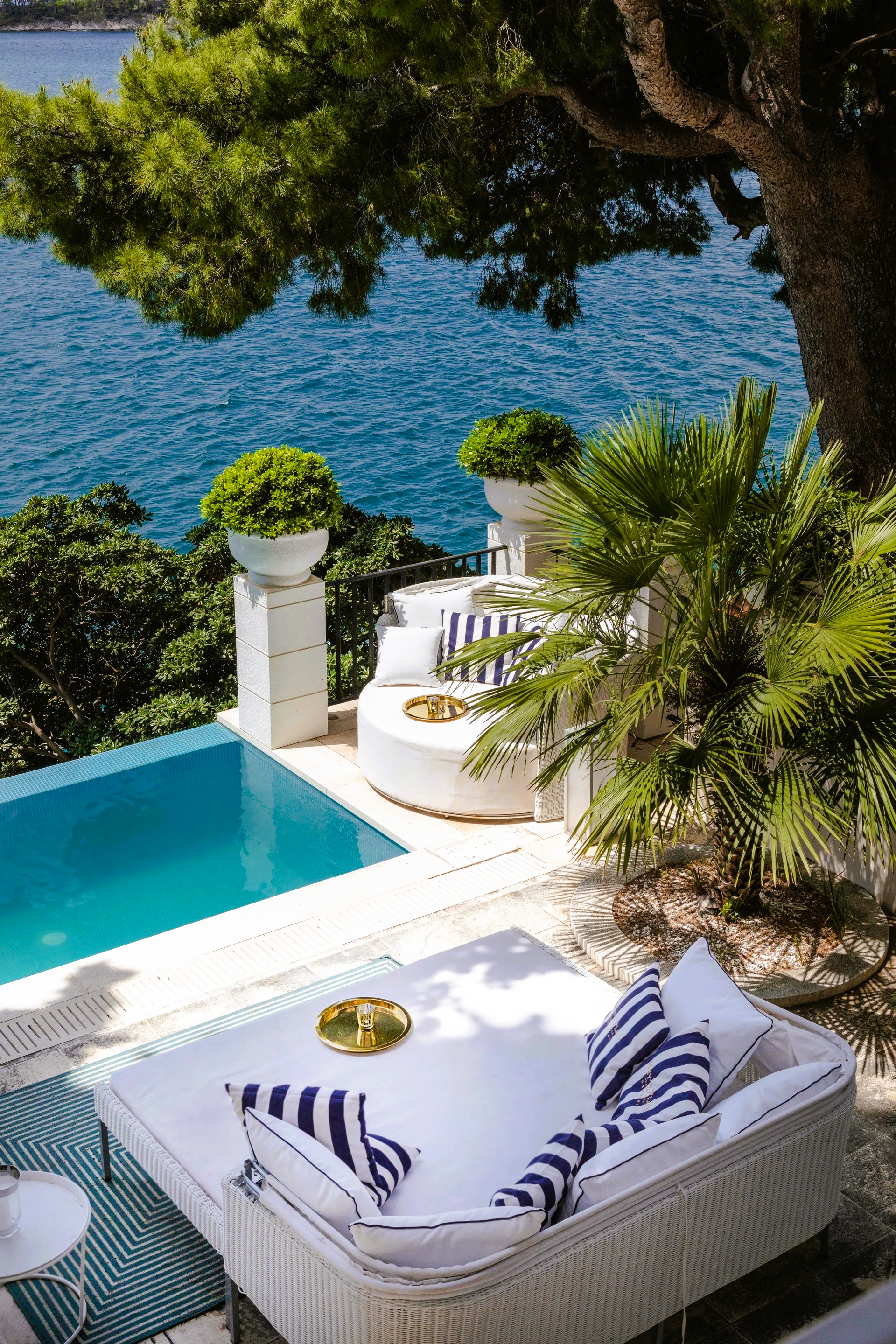 Poolside seating overlooking turquoise water. White lounge, blue striped pillows, green foliage.
