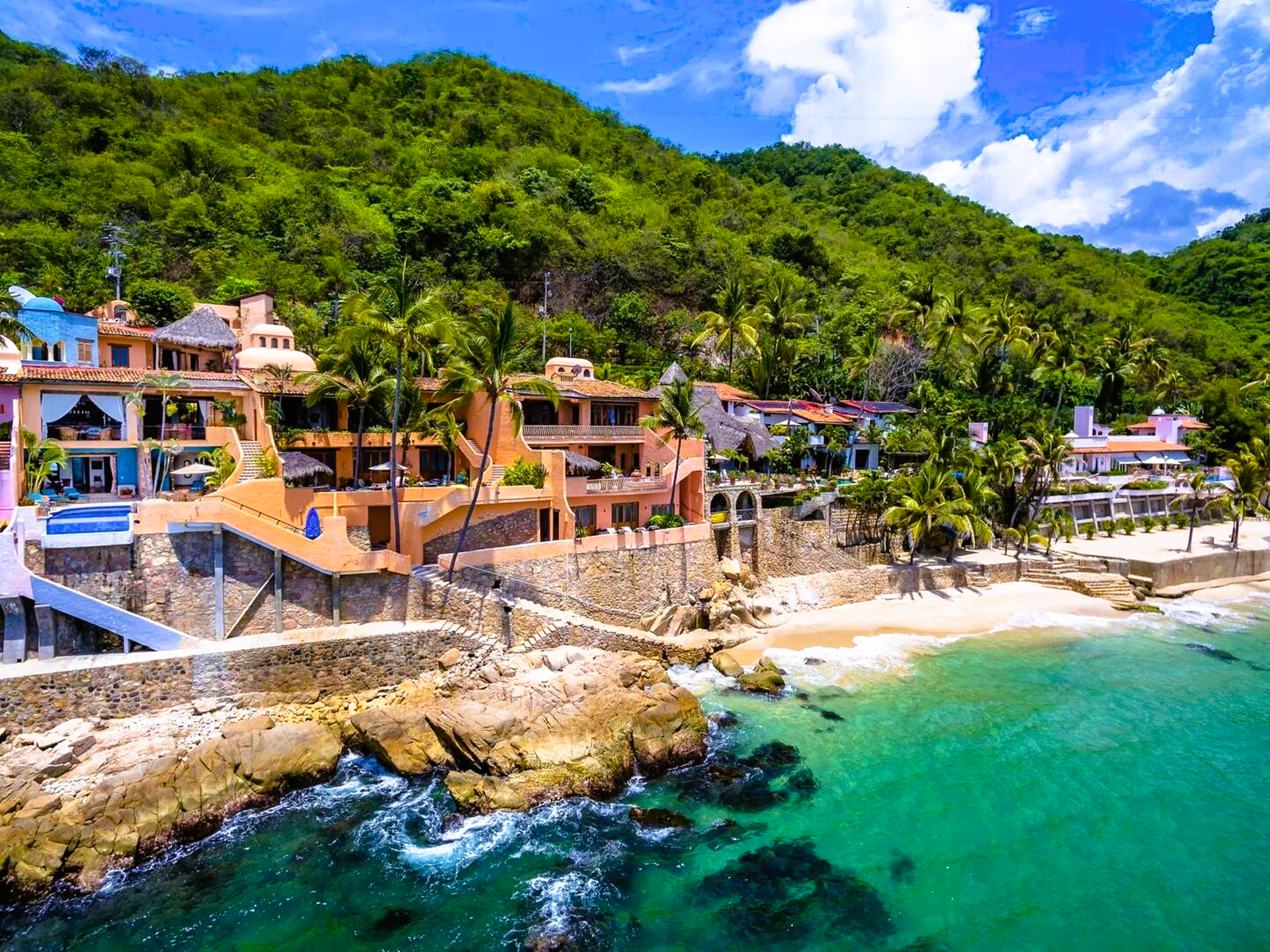 Coastal villas nestled on a rocky beach, green mountains in the background, turquoise waters.