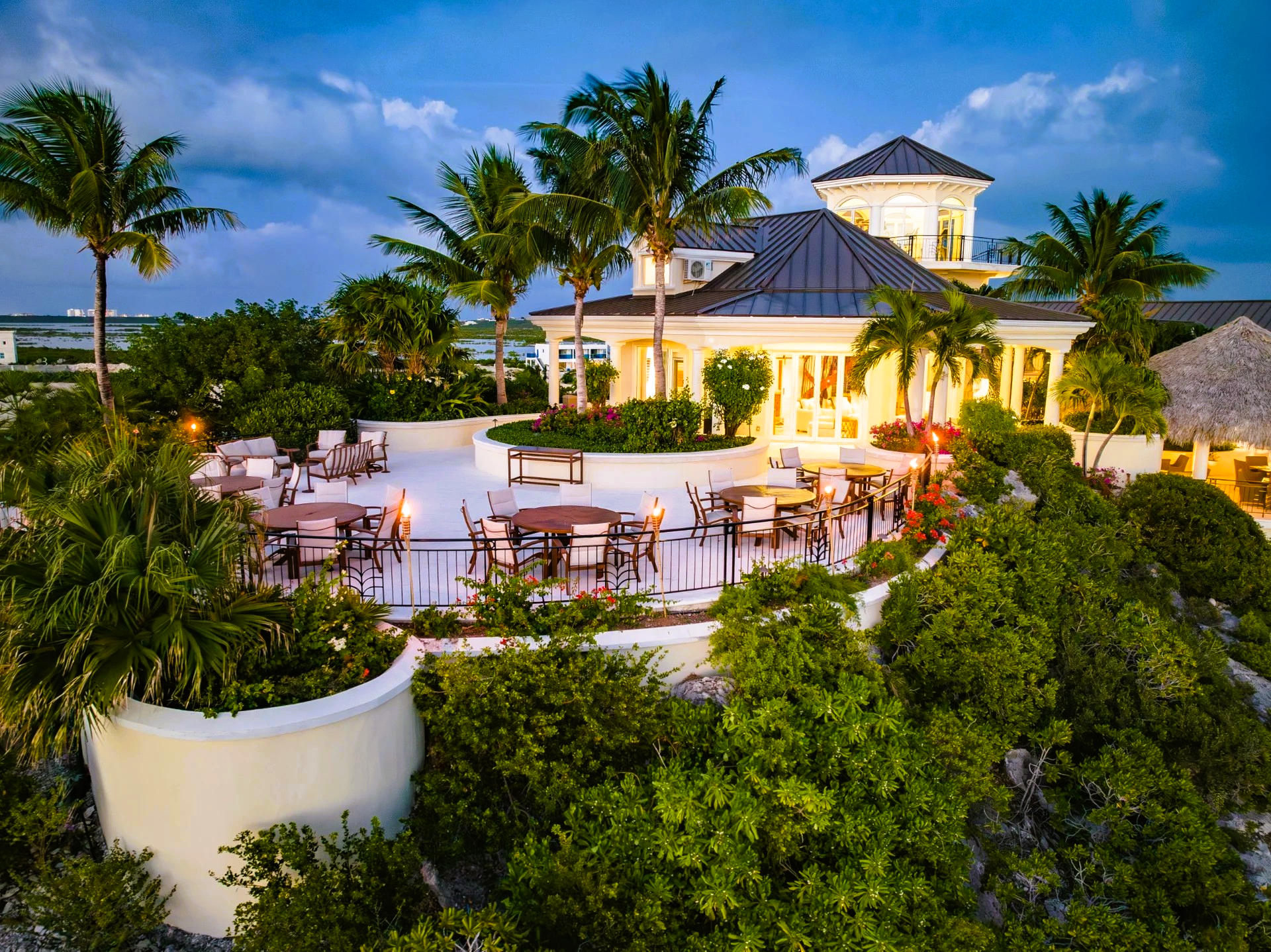 Terrace restaurant at dusk with tables, lush greenery, and palm trees.