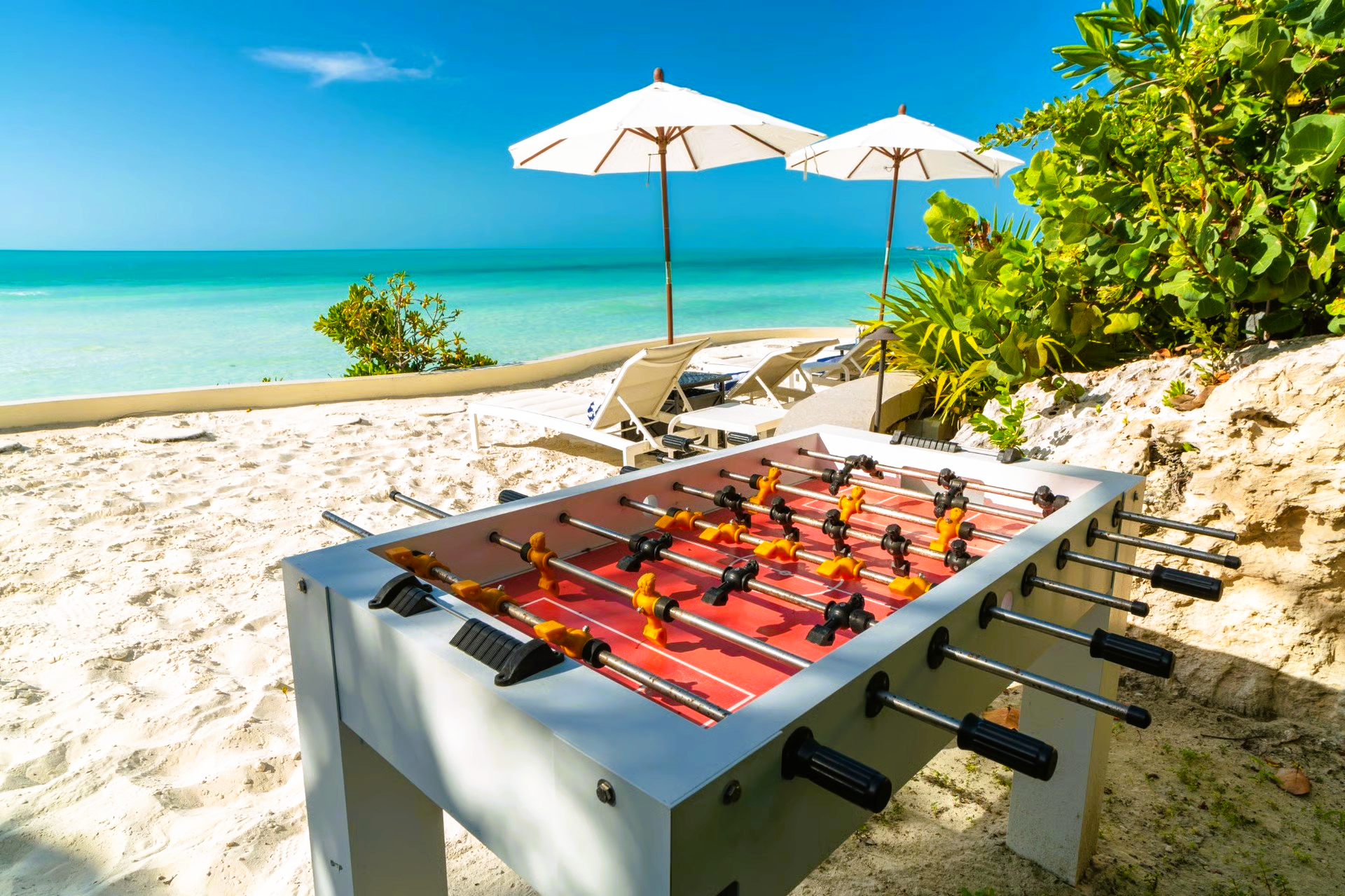 Foosball table on a sandy beach with umbrellas and turquoise water in the background.
