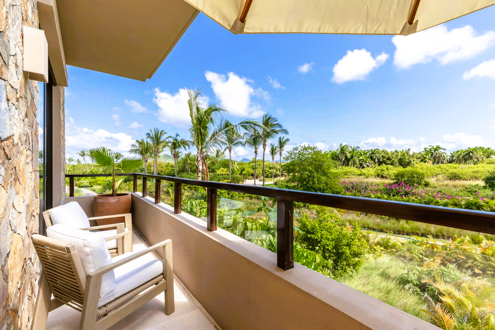 Balcony overlooking lush green landscape and blue sky; two chairs, beige, stone wall.