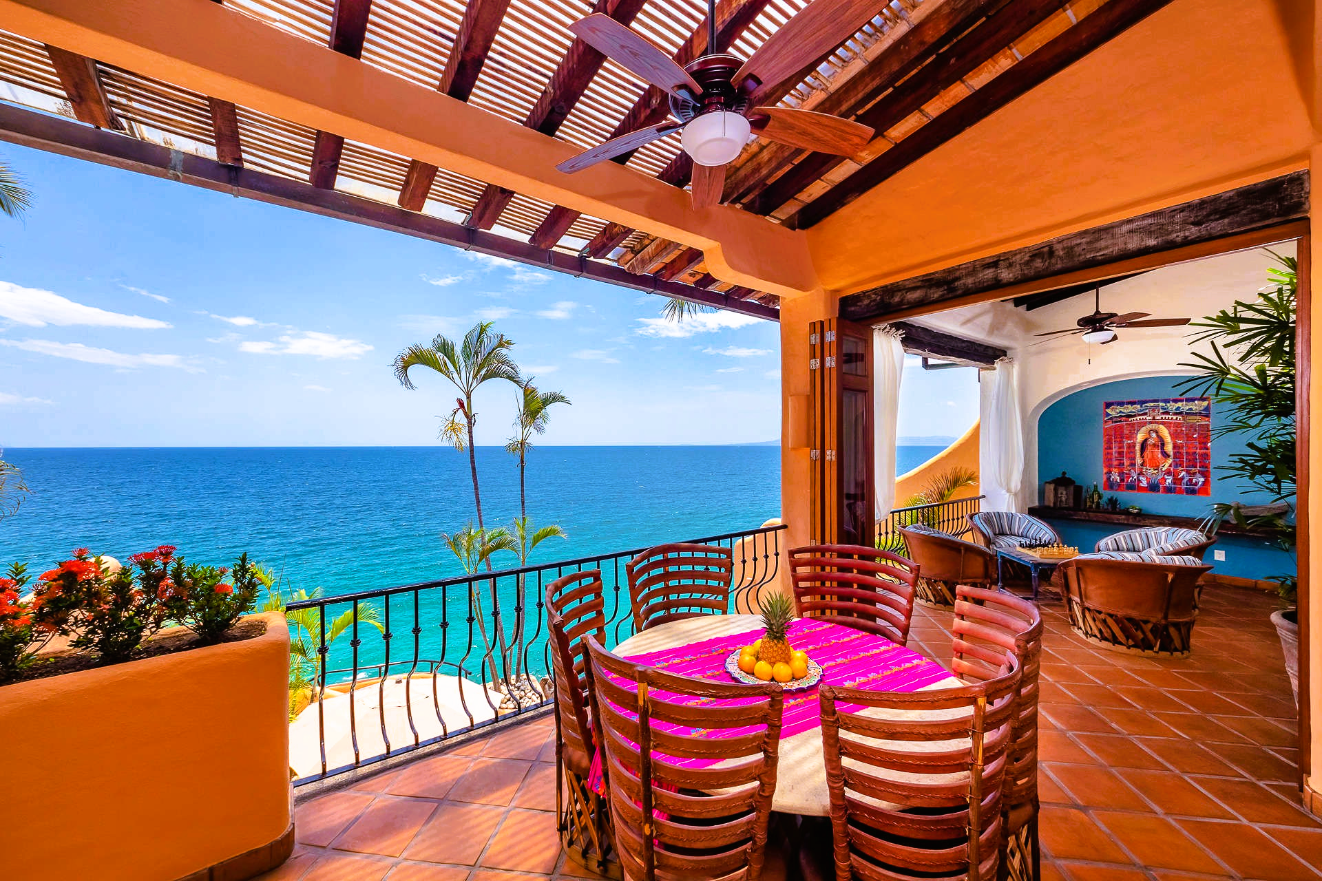 Terrace overlooking ocean with table, chairs, and plants. Orange walls, blue water, sunny day.