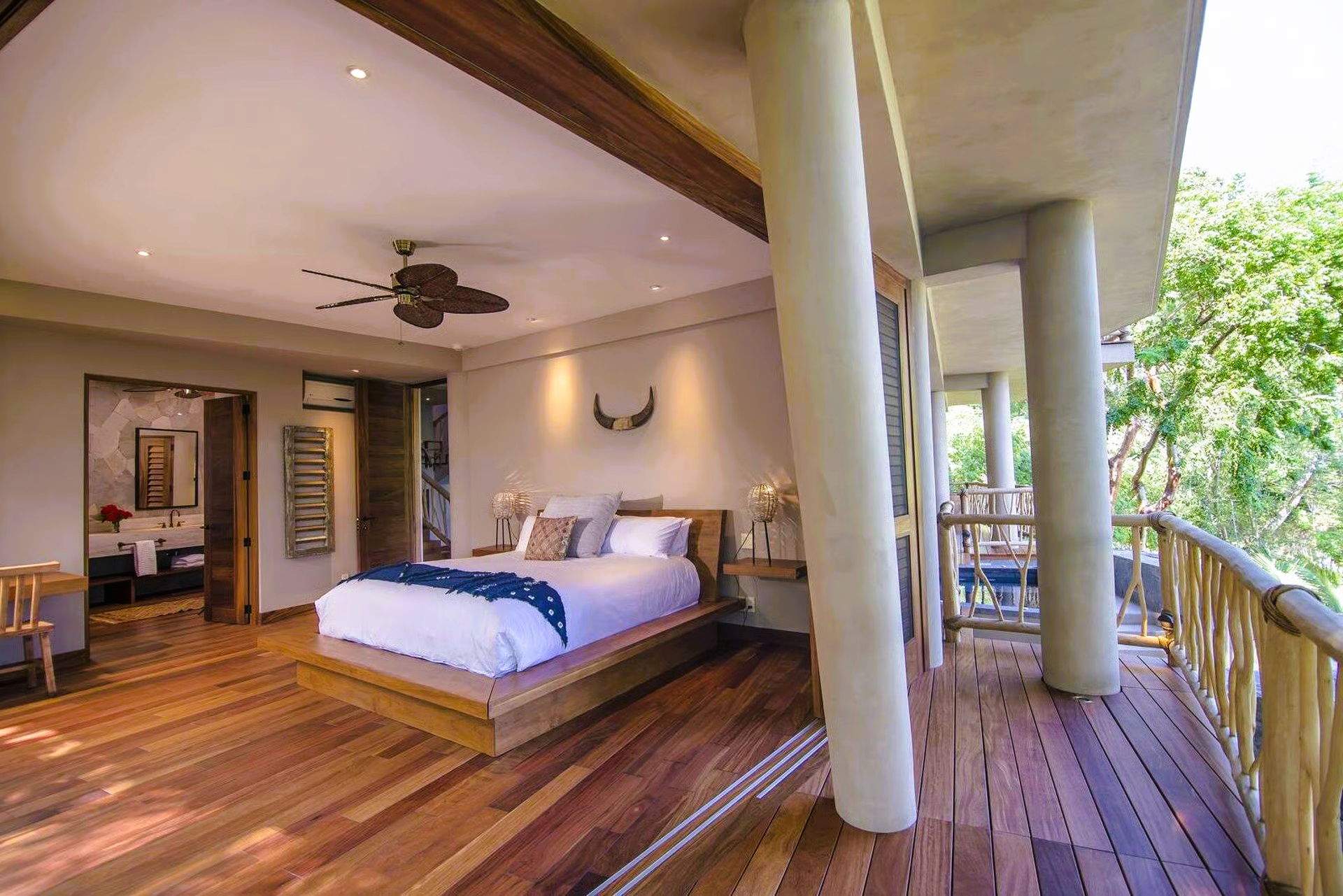 Bedroom with wood floors, bed, and balcony overlooking trees.