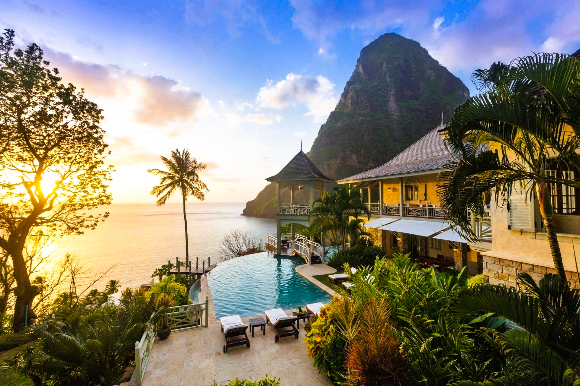 Luxury resort with a pool overlooking the ocean, palm trees, and the Pitons mountains at sunset.