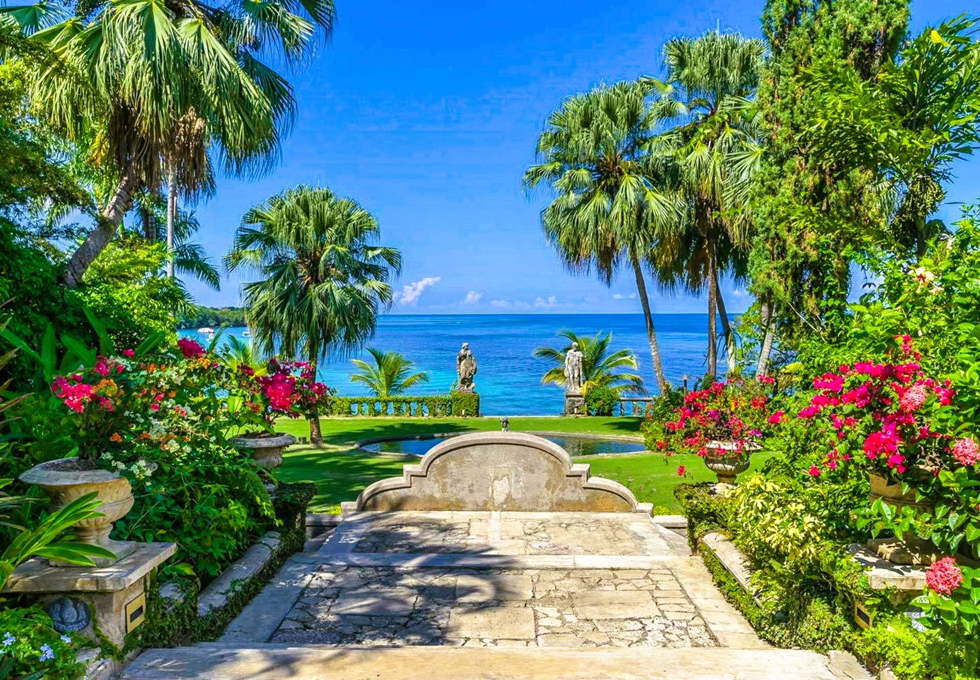 Stone steps lead to a lush garden overlooking a turquoise ocean, framed by palm trees and flowers.