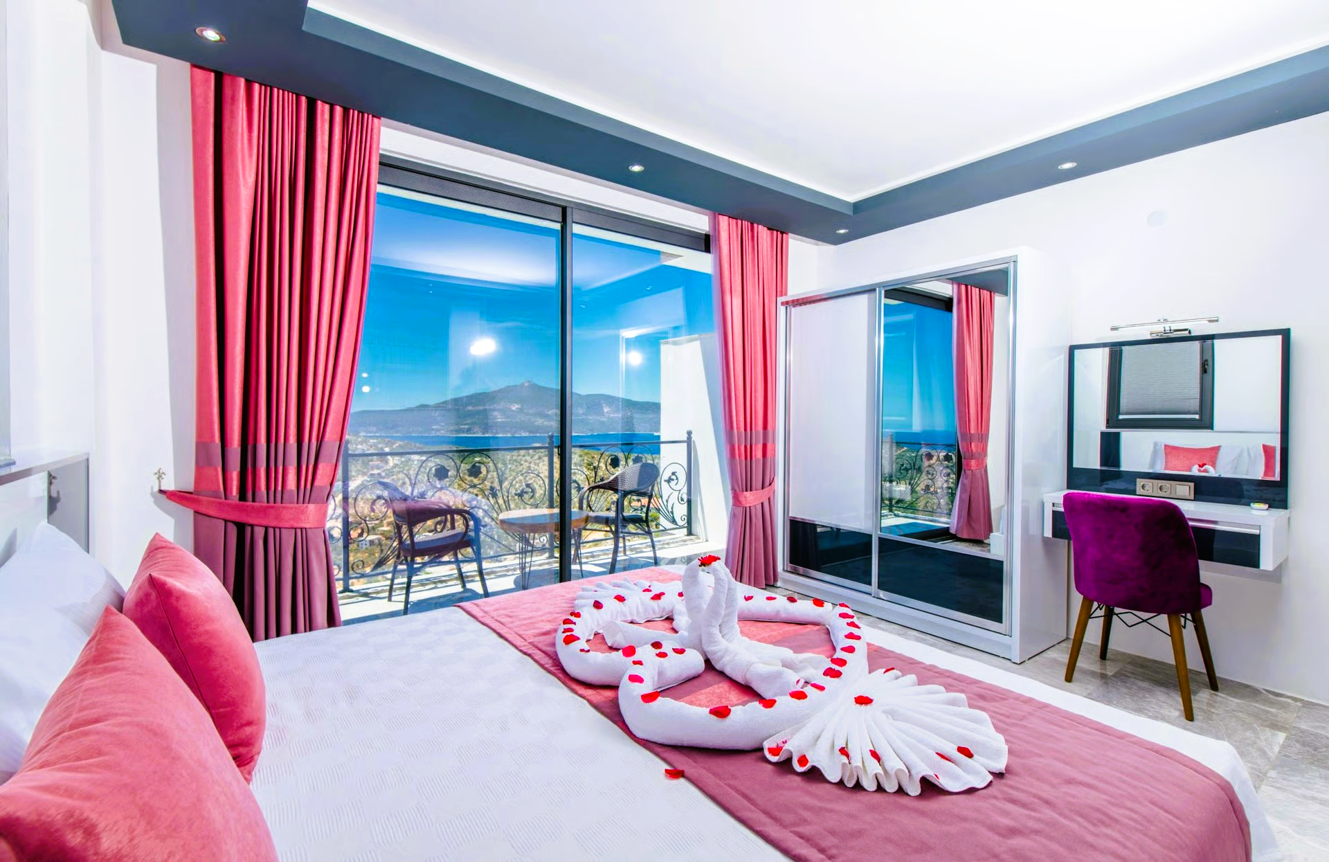 Bedroom with sea view, pink curtains, bed decorated with towel swans and roses, vanity.