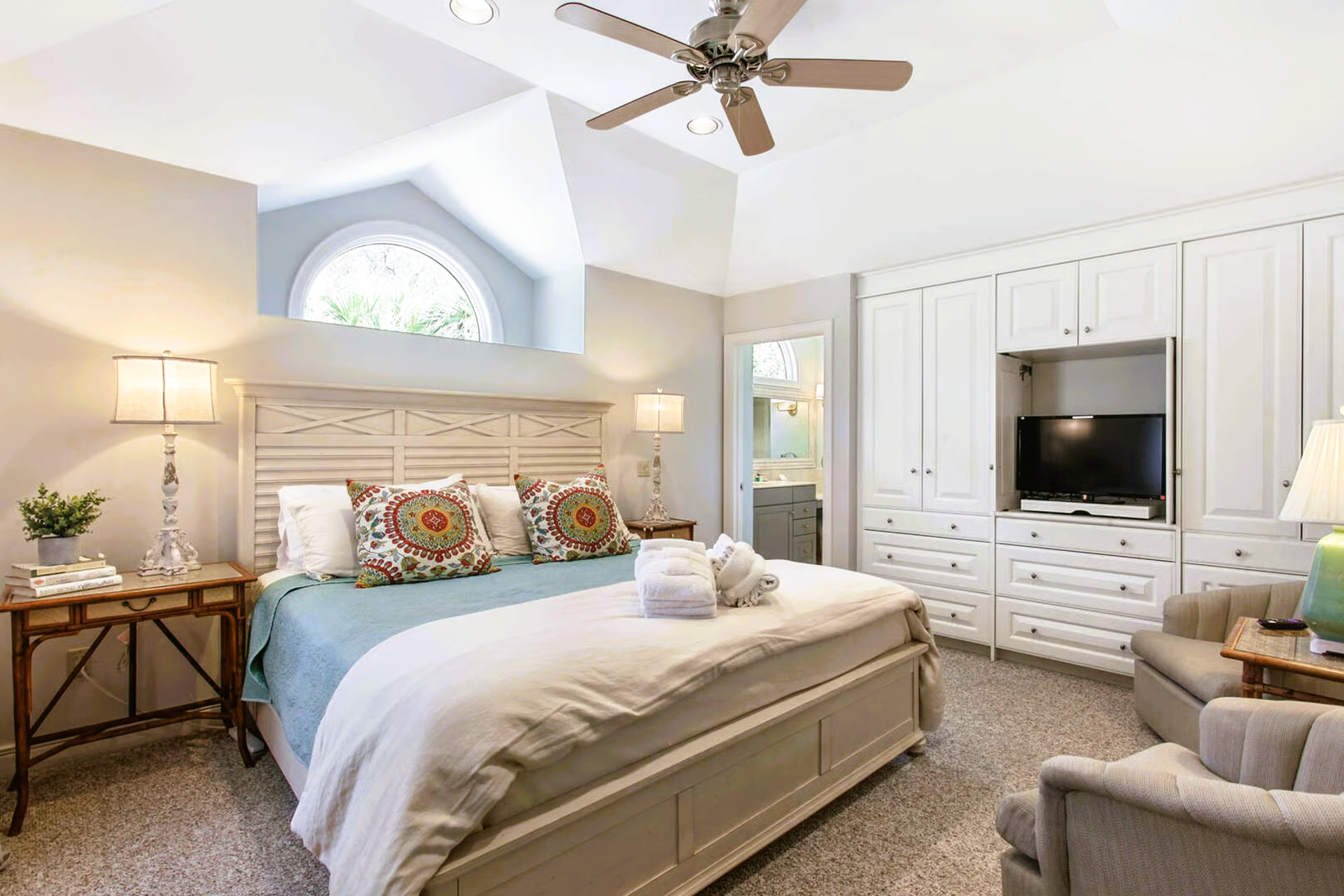Bedroom with king-size bed, built-in white cabinets, TV, two armchairs, side table, and ceiling fan.