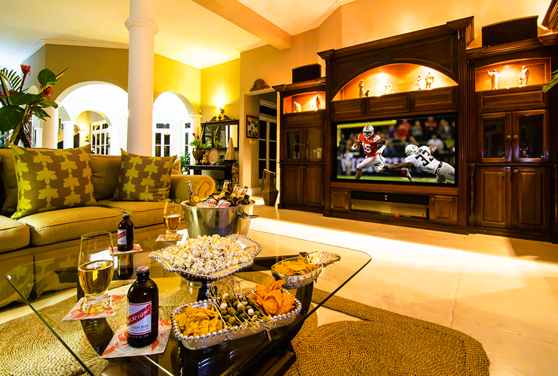 Living room with TV showing a football game, snacks on coffee table, drinks, and a gold sofa.