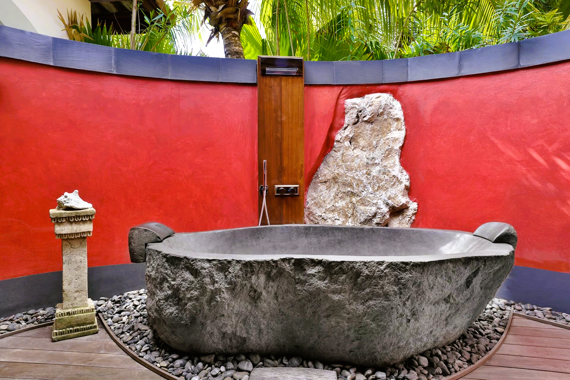 Stone outdoor bathtub set in a red-walled garden with a stone sculpture and a small statue.