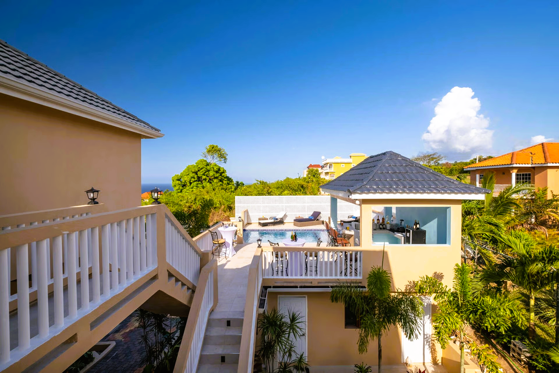 Exterior view of a beige house with a pool, stairs, and blue sky. Palm trees and other buildings surround the property.